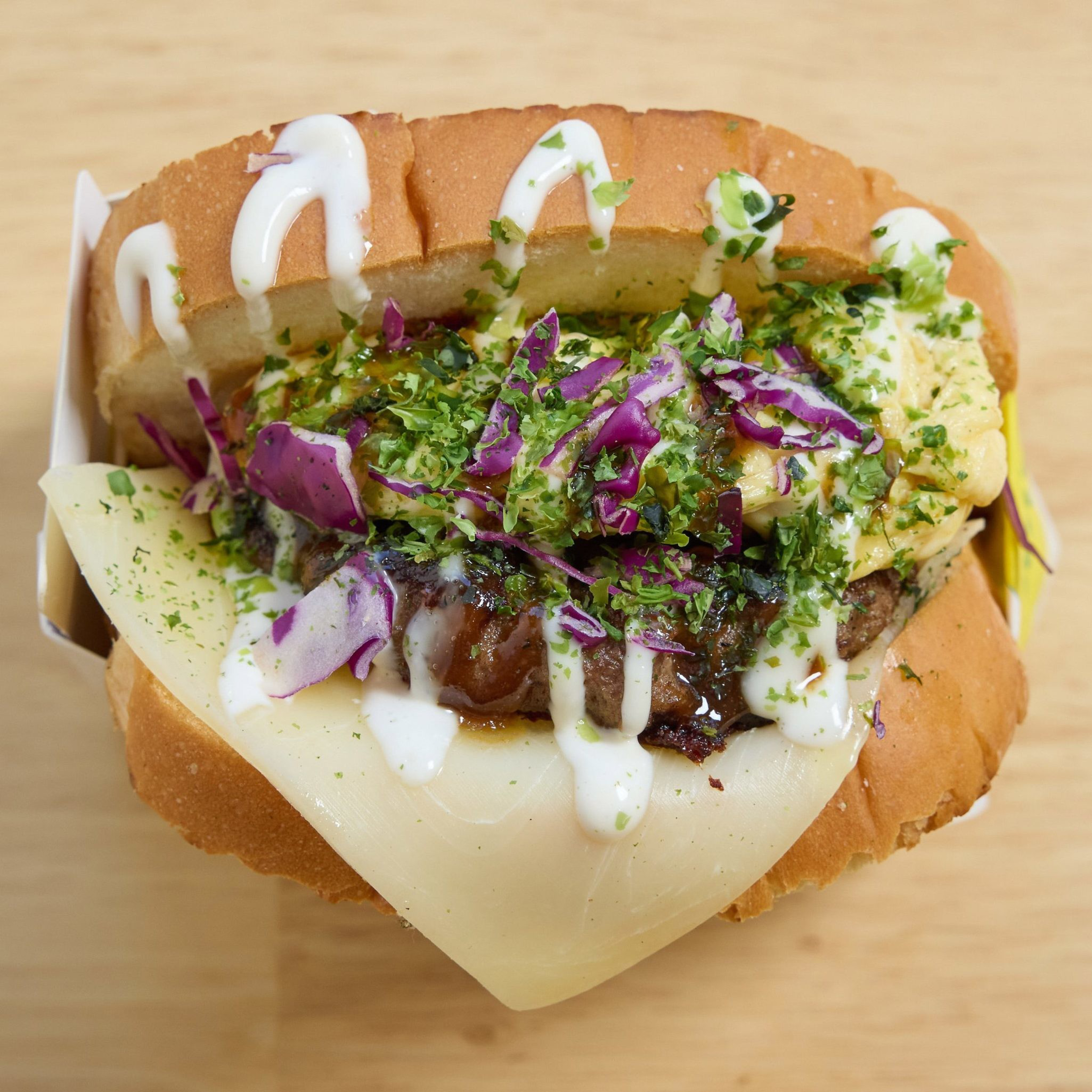 The image shows a hot dog topped with melted cheese, chopped purple cabbage, drizzled white sauce, and a green herb garnish, set on a light wooden surface.
