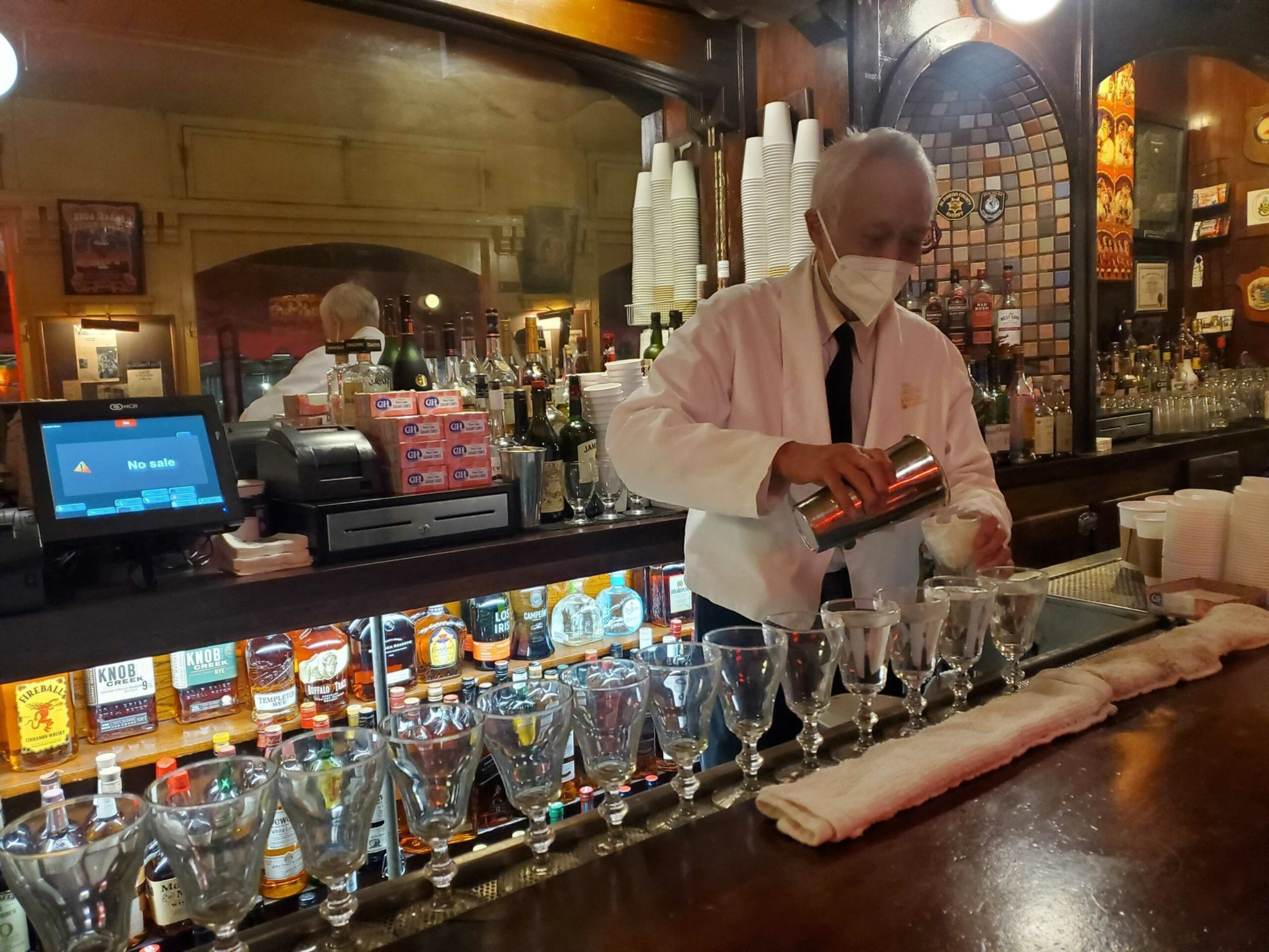 A masked bartender in a white jacket pours a drink from a shaker into a glass on a bar lined with other empty glasses.
