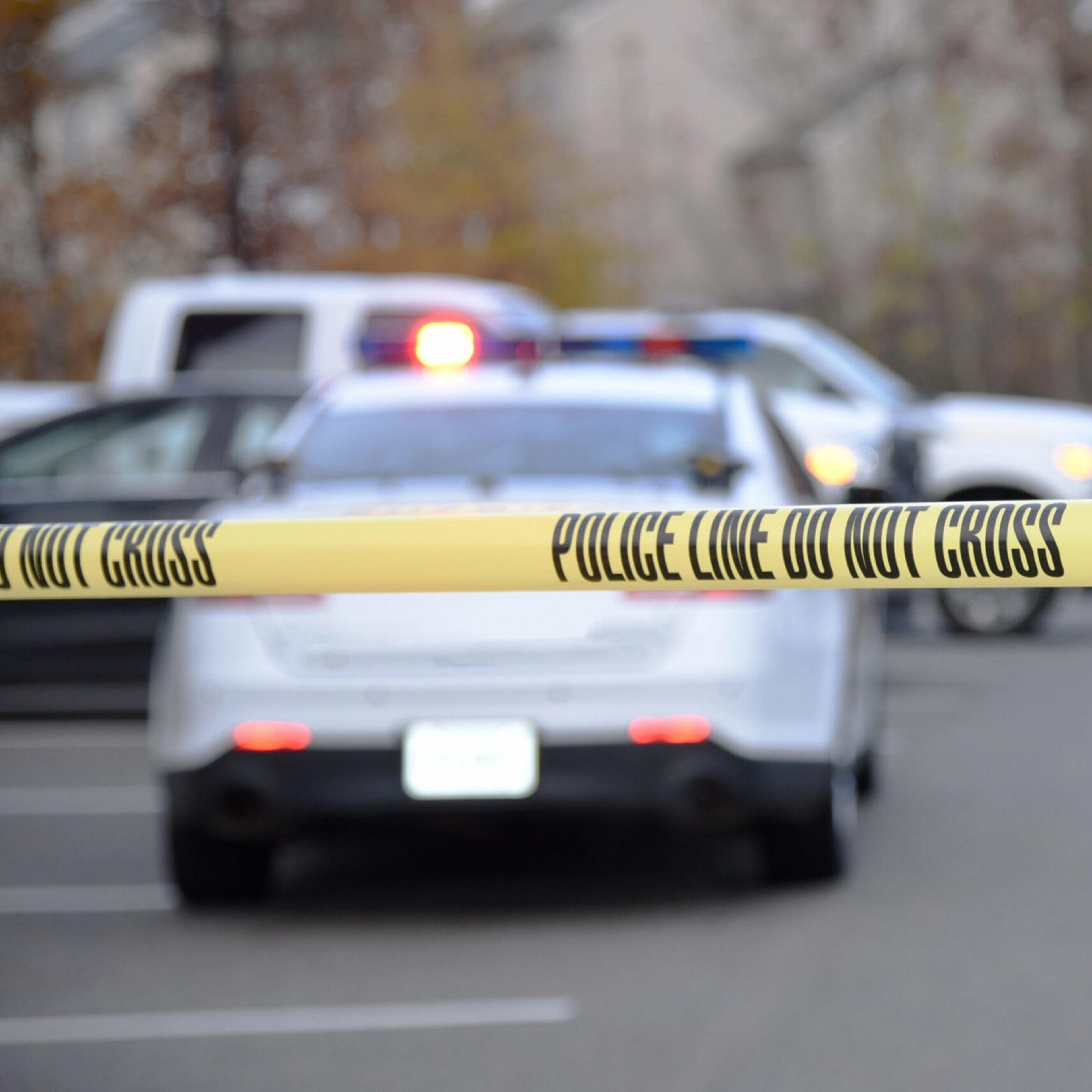 Police tape is seen close up, with a police patrol vehicle some distance beyond.