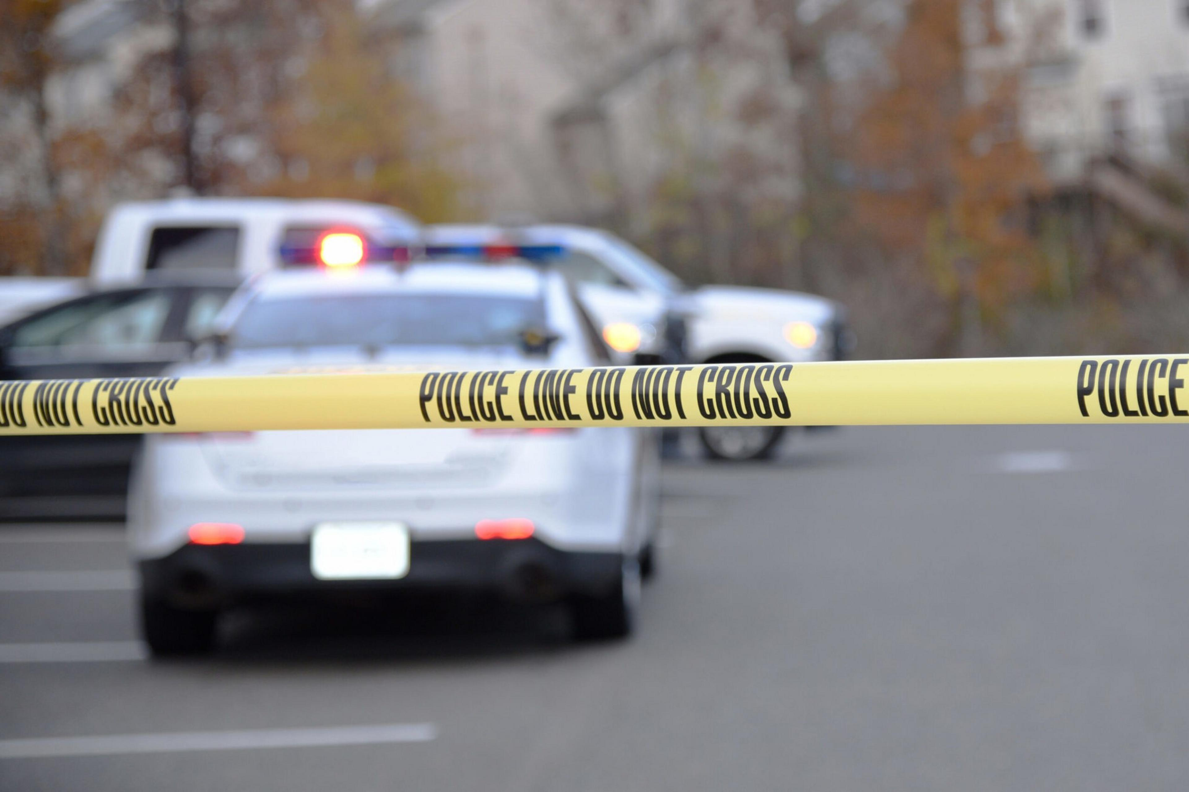 Police tape is seen close up, with a police patrol vehicle some distance beyond.