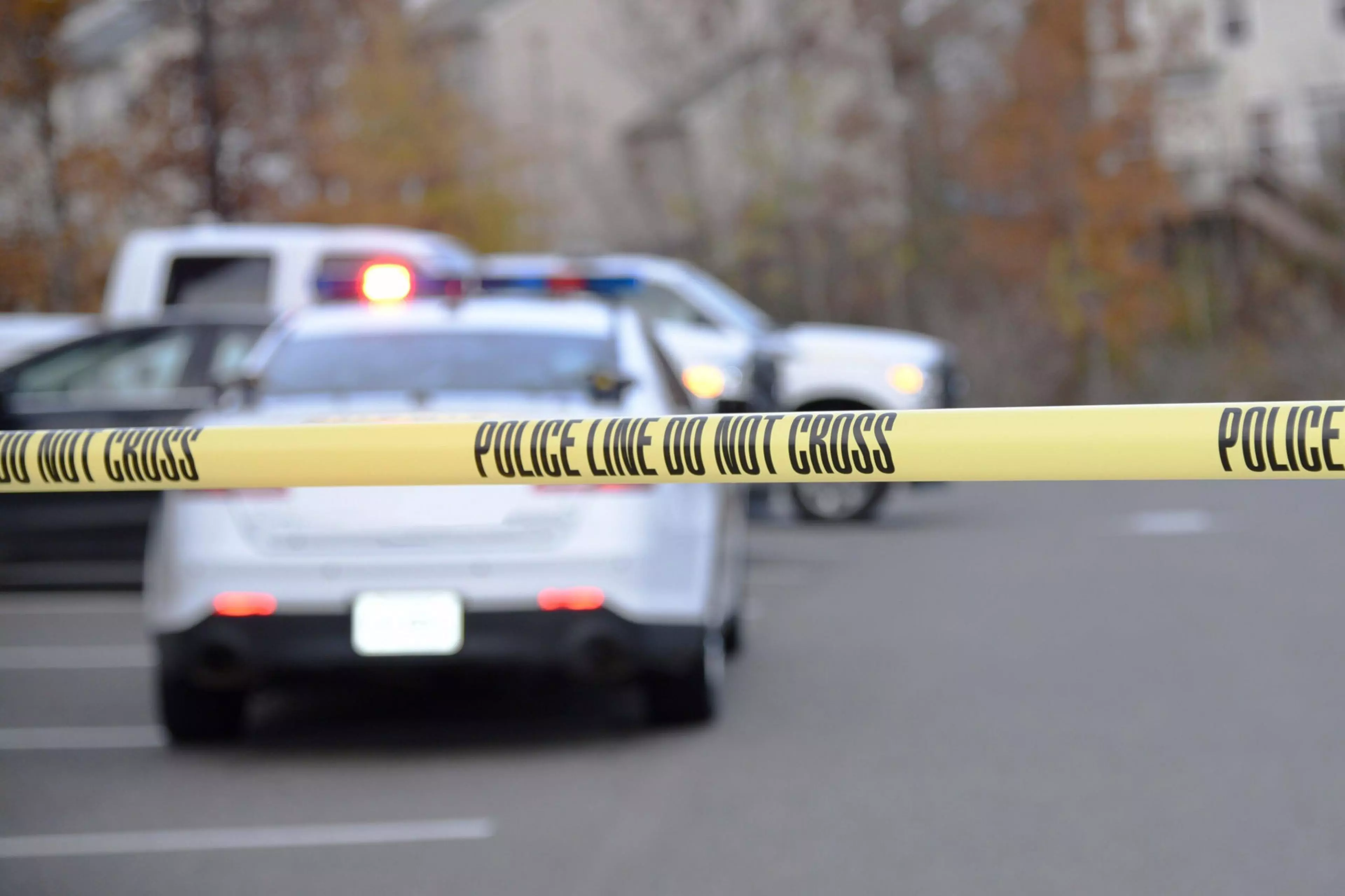 Police tape is seen close up, with a police patrol vehicle some distance beyond.