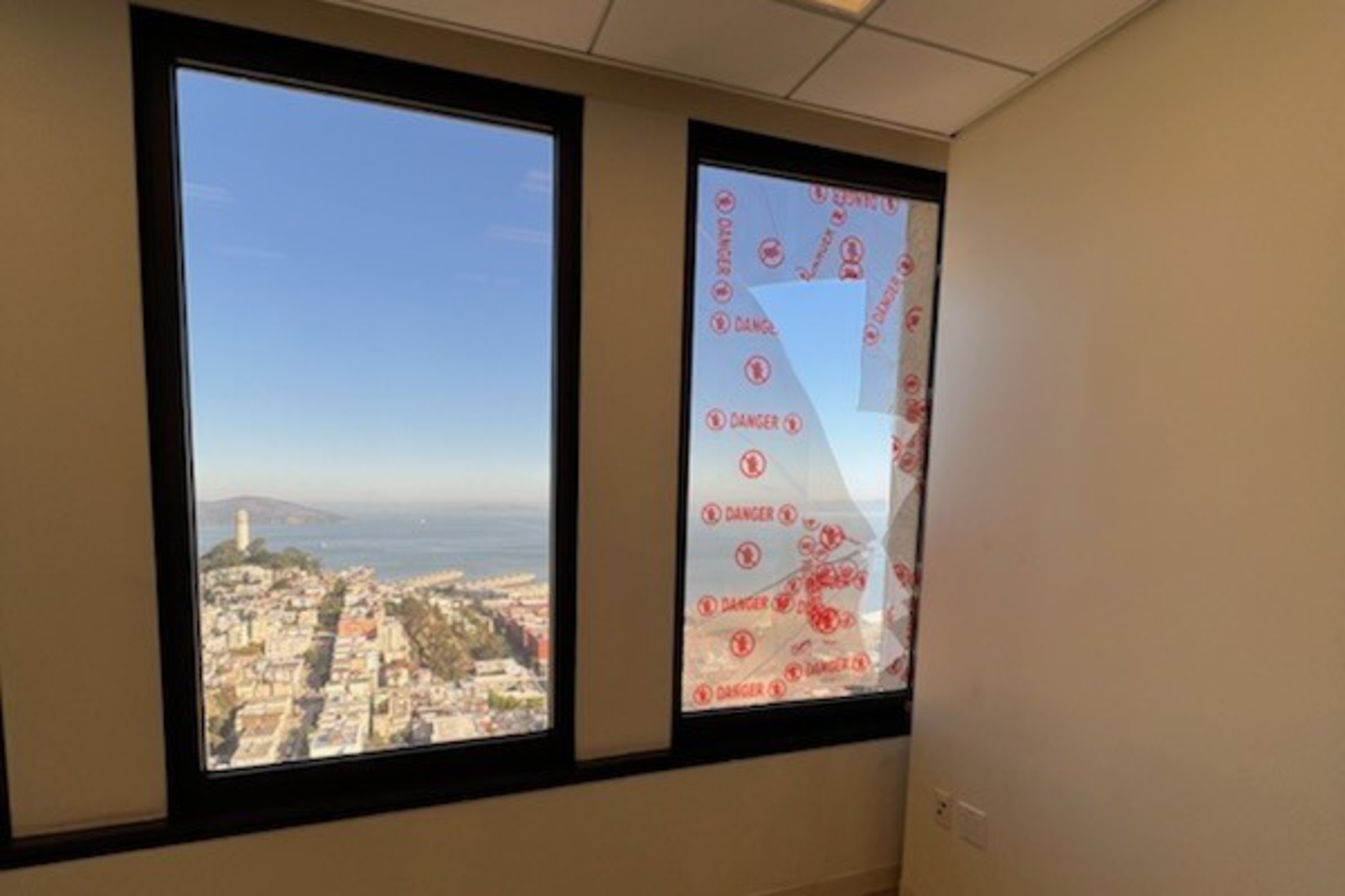 The image shows a room with two large windows; one has a clear view of a cityscape and the ocean, while the other is covered in red "DANGER" stickers.