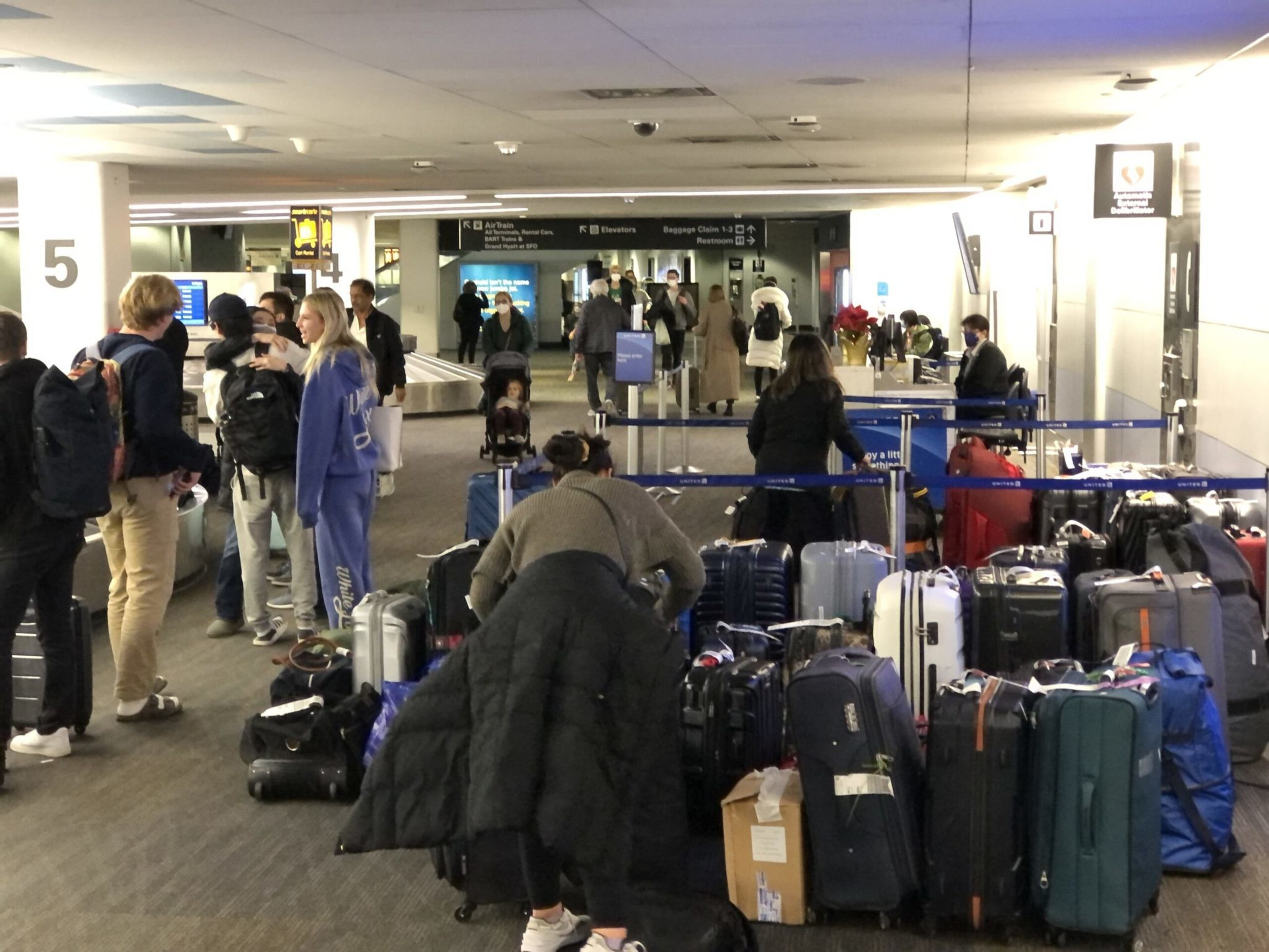 Several people stand and wait near baggage claim at an airport, with many suitcases and bags piled in the foreground and a few travelers walking in the background.