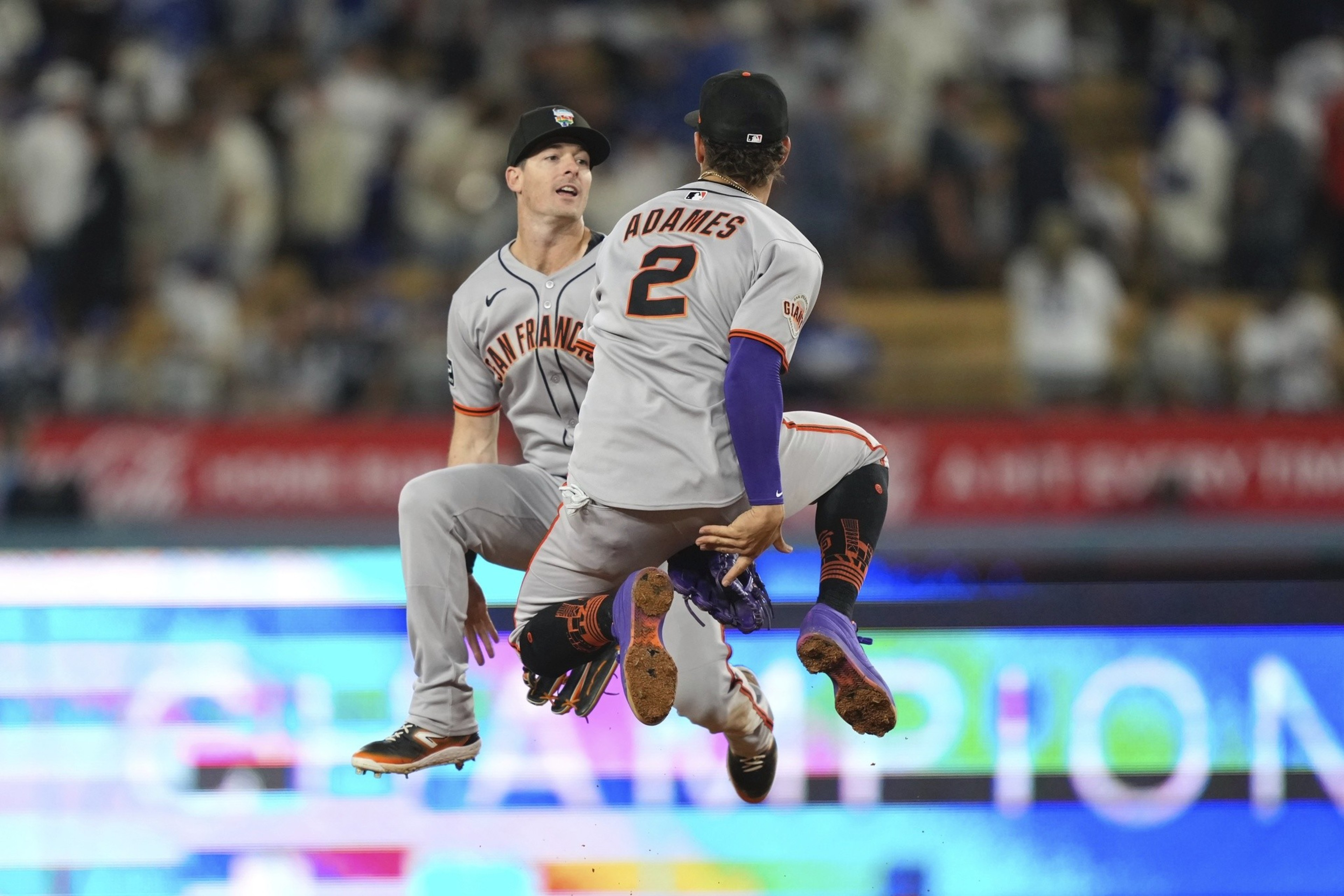 Two baseball players in gray uniforms are mid-air, celebrating. One player's back is visible with &quot;ADAMES&quot; and number 2. They're on a baseball field with a blurred crowd.