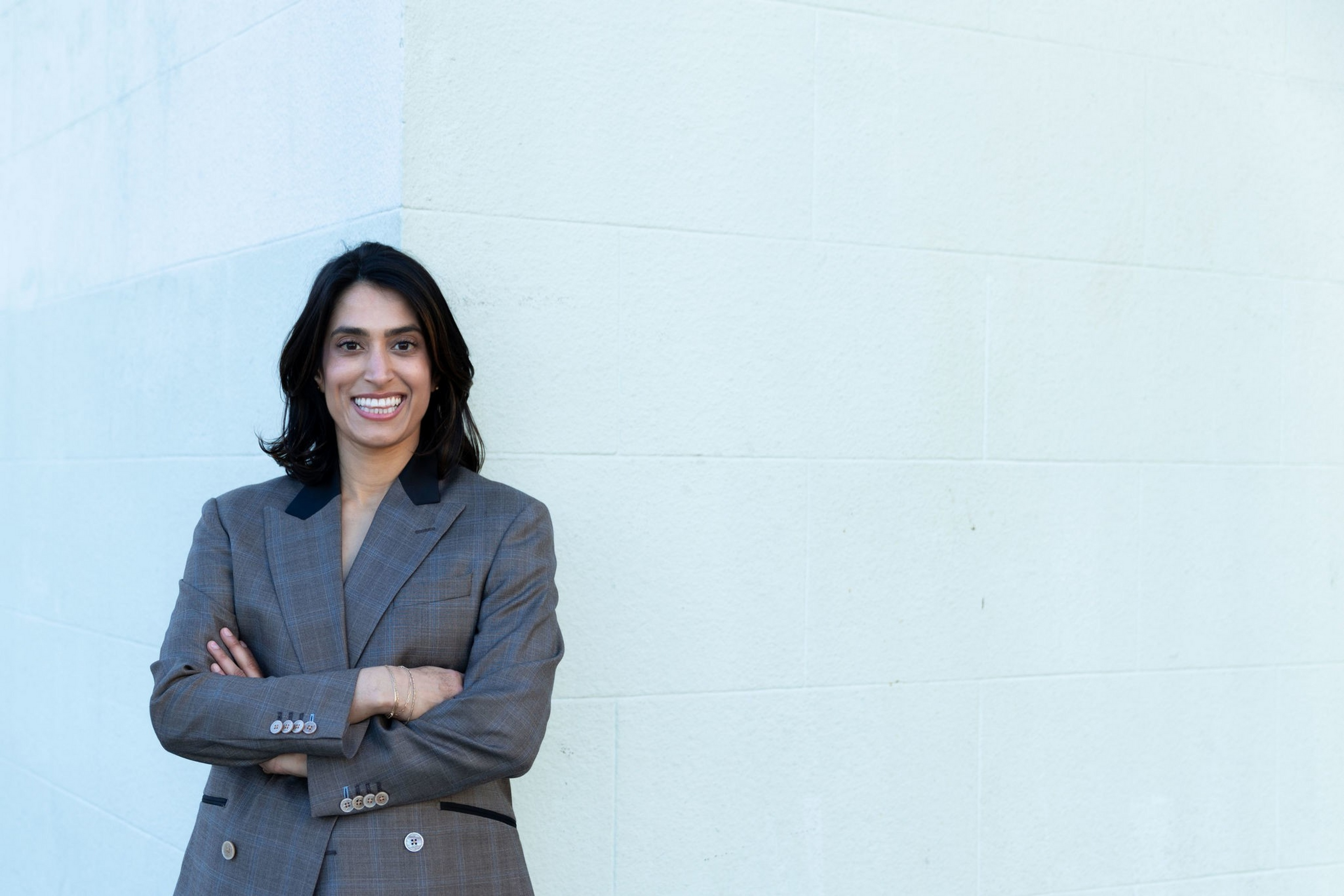 A smiling woman with shoulder-length dark hair stands with arms crossed wearing a gray blazer against a plain light-colored wall.