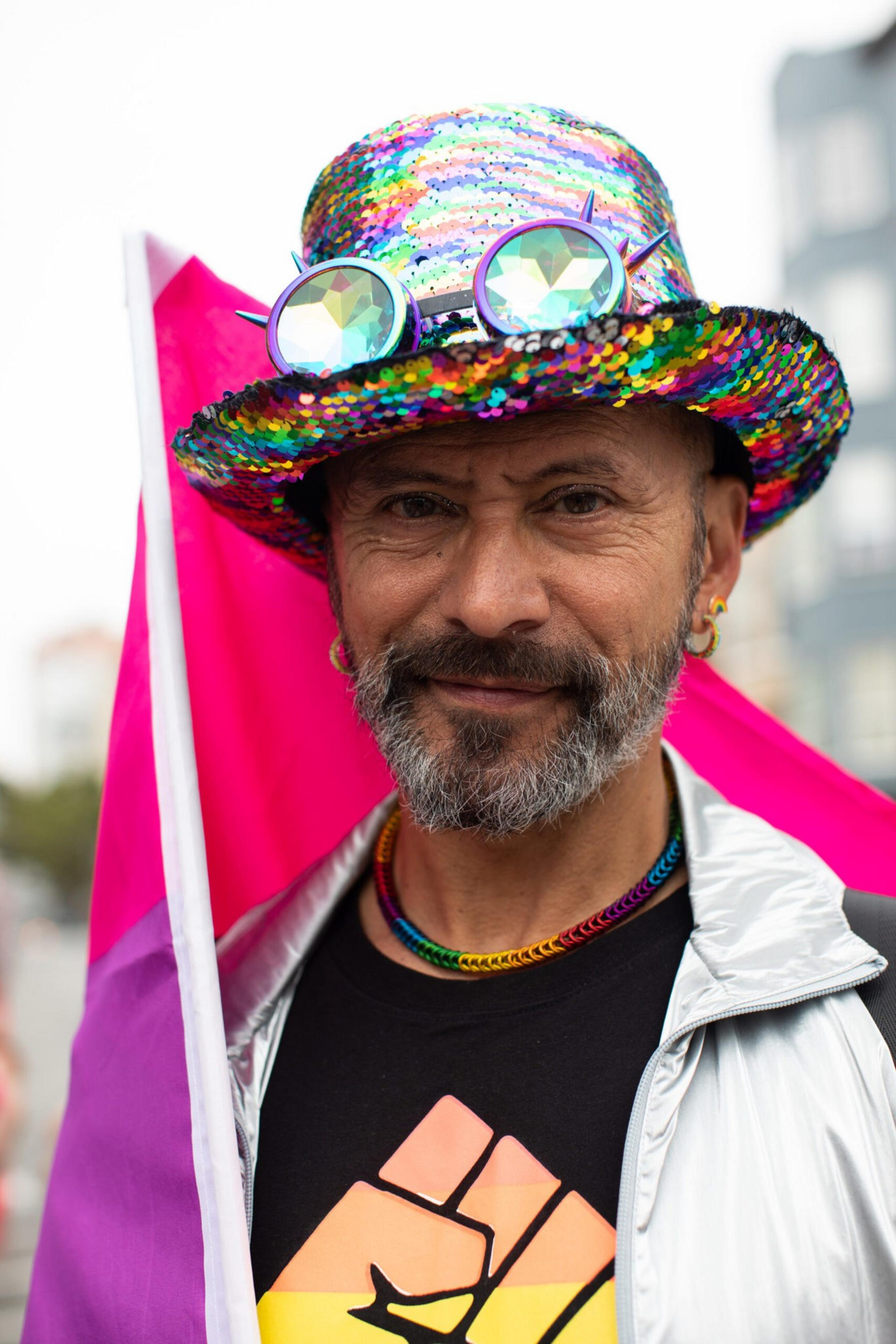 A man wears a multicolored sequin hat with large round glasses, a rainbow necklace, and a black shirt with a raised fist symbol while holding a pink and purple flag.