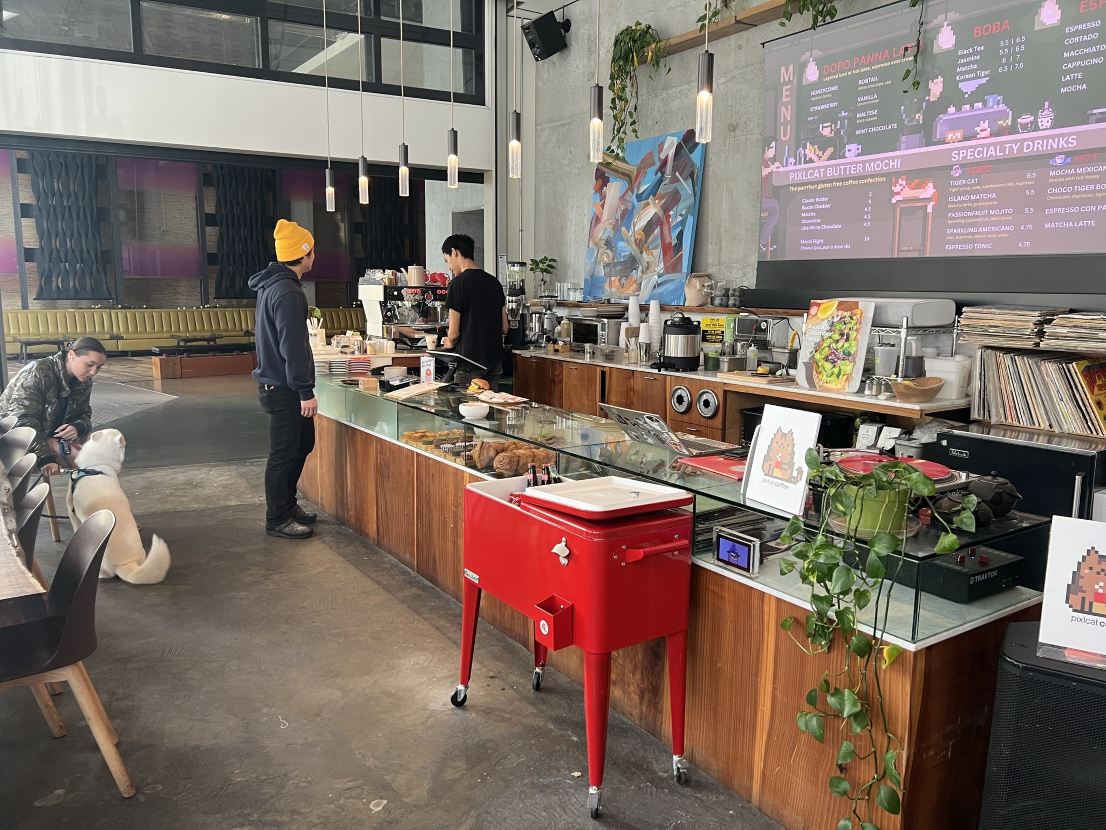 A long wooden bar stands inside a coffee shop with a menu screen behind it. 