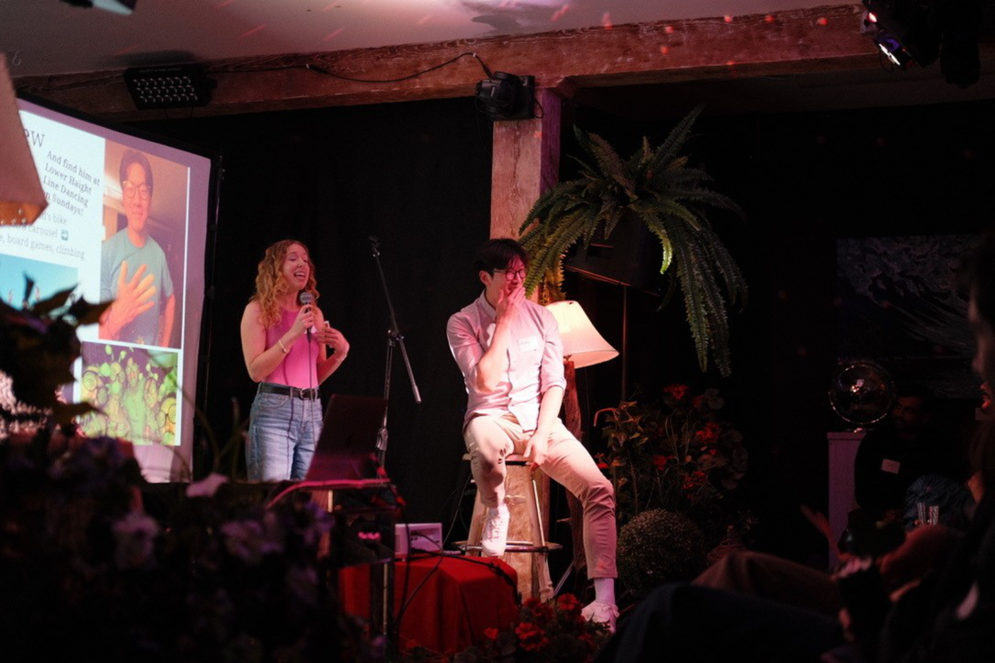 A woman stands smiling with a microphone, and a man sits on a stool beside her, partially covering his face with his hand, in a warmly lit room.