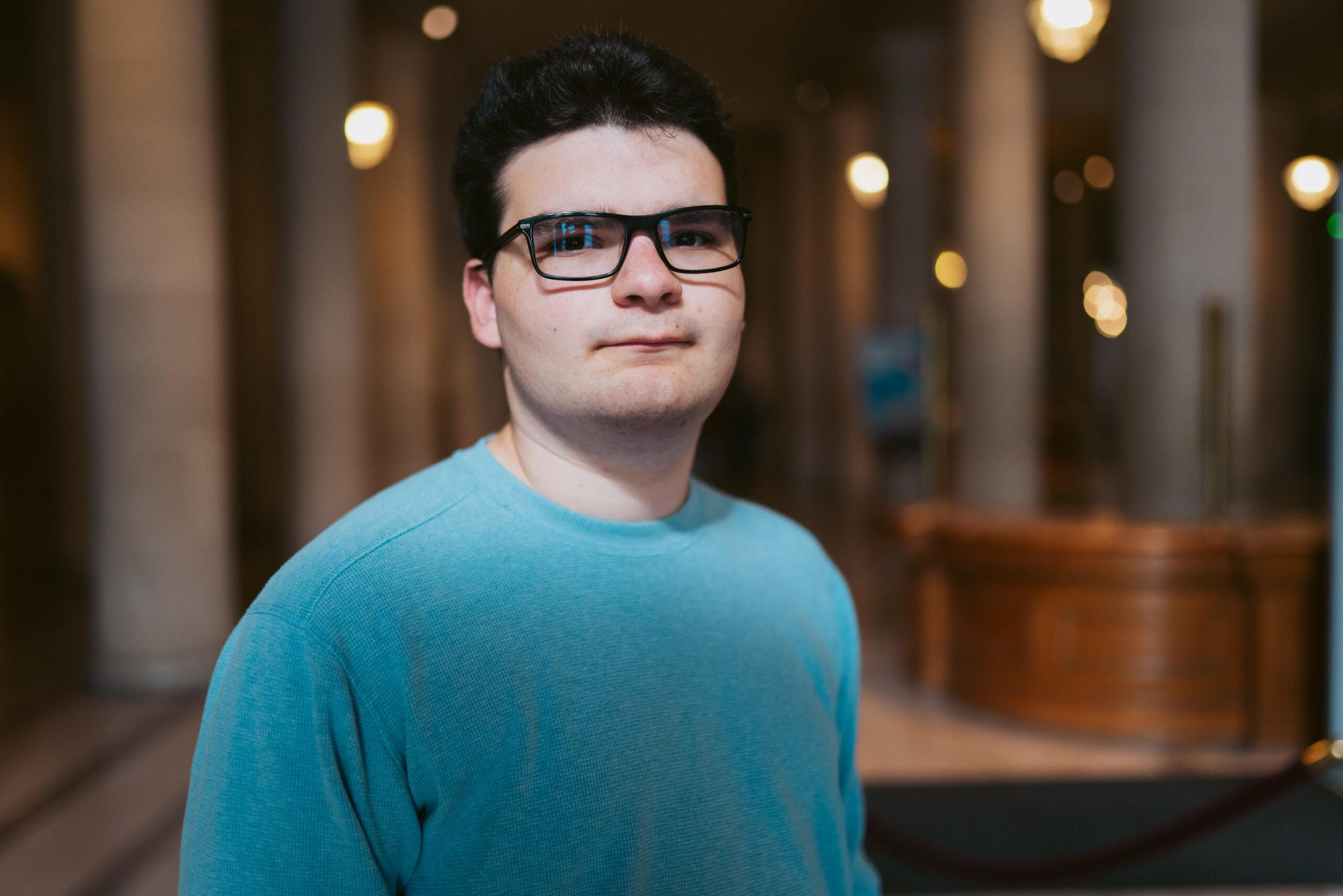 A young man with short dark hair and glasses wears a light blue sweater and stands indoors in a softly lit space with tall pillars and blurred background lights.