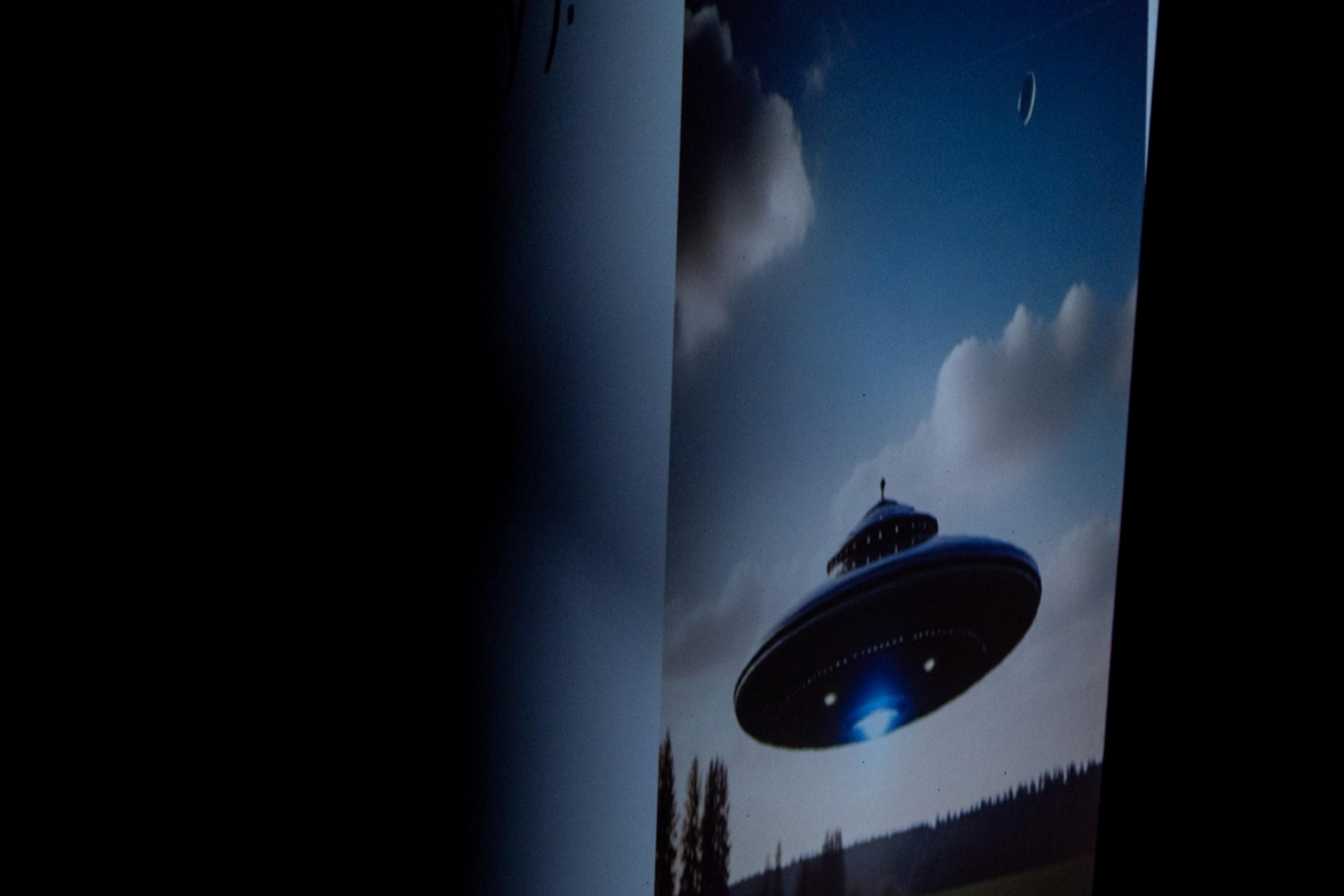 The image shows a UFO hovering over a grassy field with trees and a partly cloudy sky in the background. There's a small moon visible in the sky.