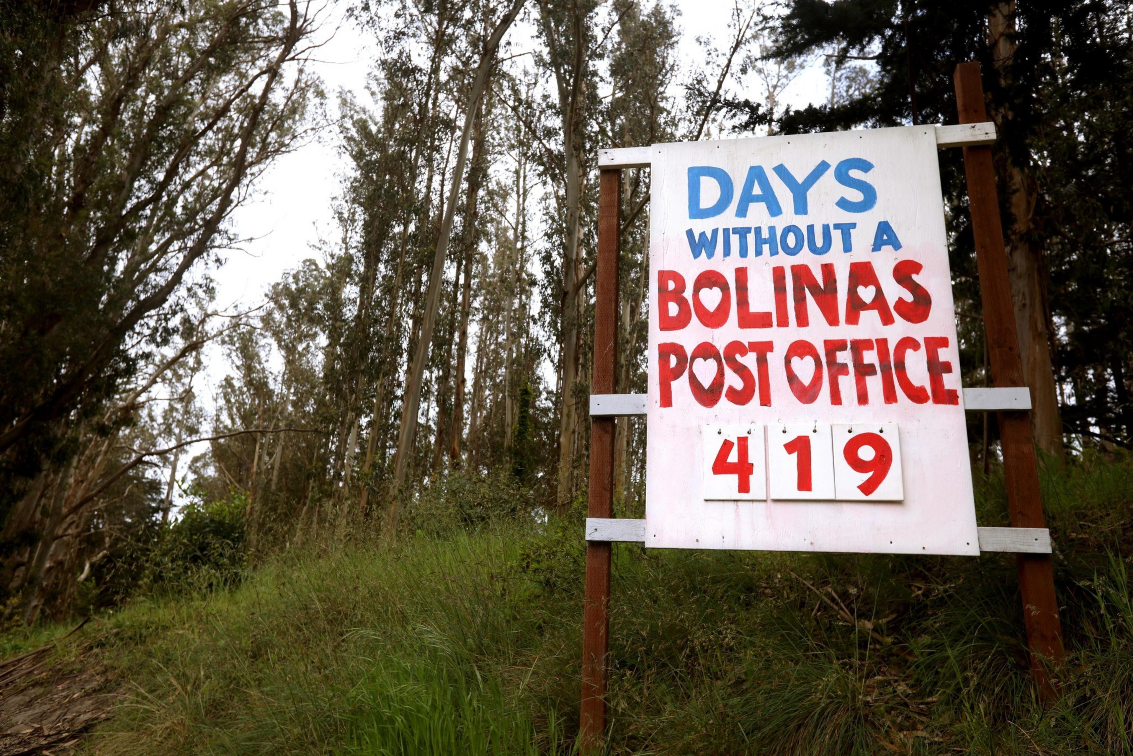 A sign stands in a wooded area, reading "DAYS WITHOUT A BOLINAS POST OFFICE" with the number "419" beneath it in bold red and white.