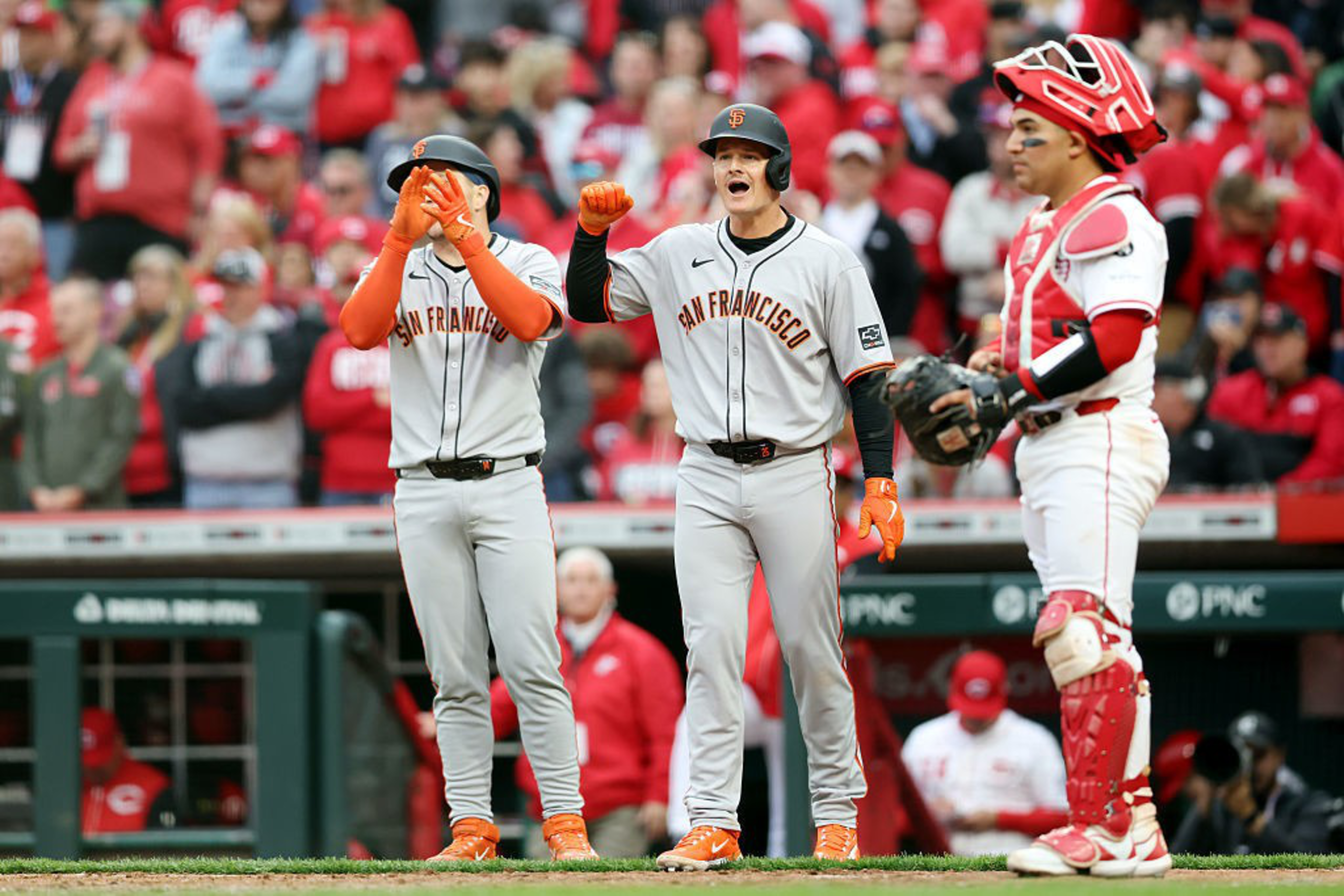 Two baseball players in grey &quot;San Francisco&quot; jerseys celebrate, with one covering his mouth. A catcher in red gear stands nearby, and fans fill the background.