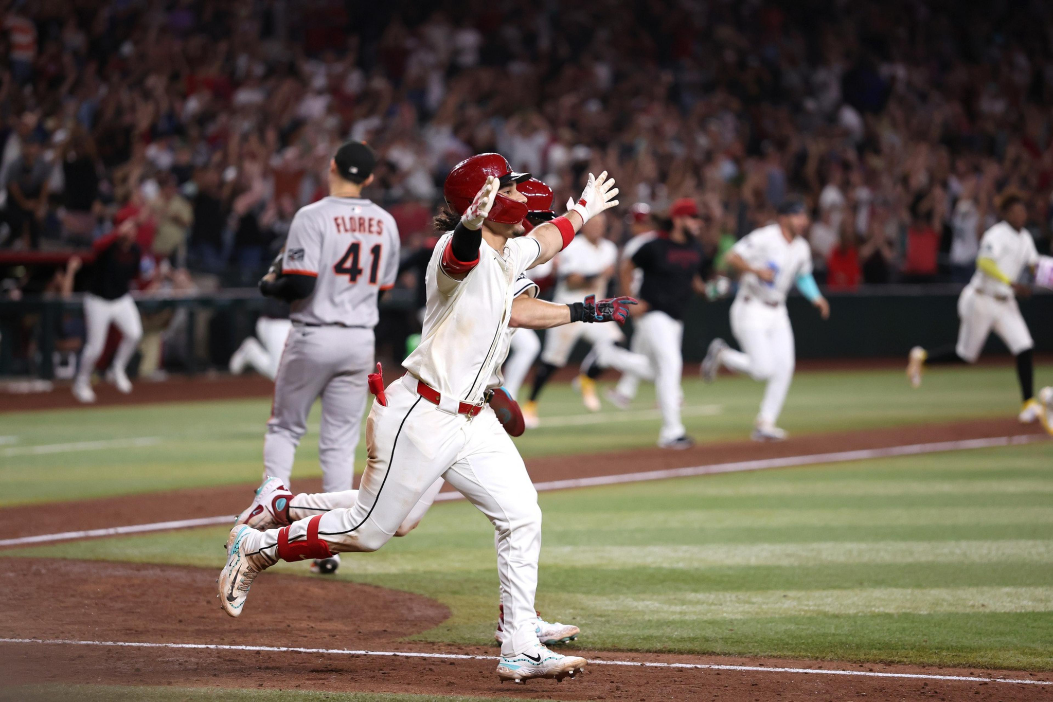 A baseball player in a white uniform celebrates with arms wide as teammates and fans cheer, while an opposing player walks away on the field.