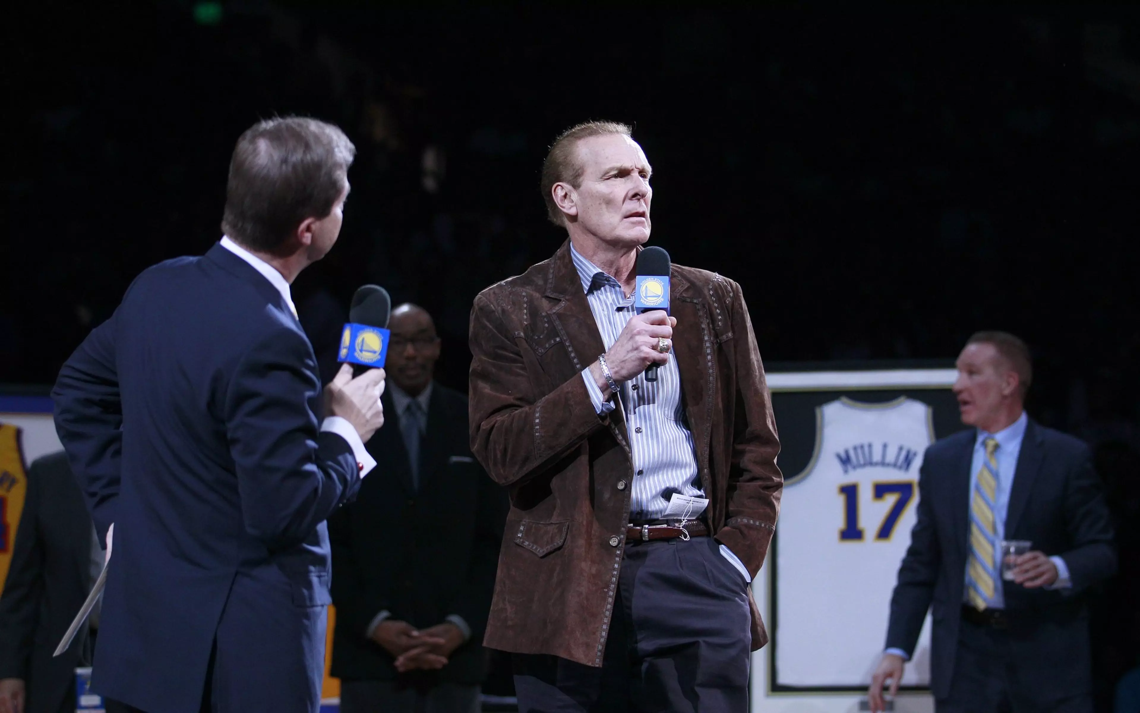 A man in a brown jacket holds a microphone speaking, while another man in a suit also holds a microphone; framed jerseys are displayed in the background.