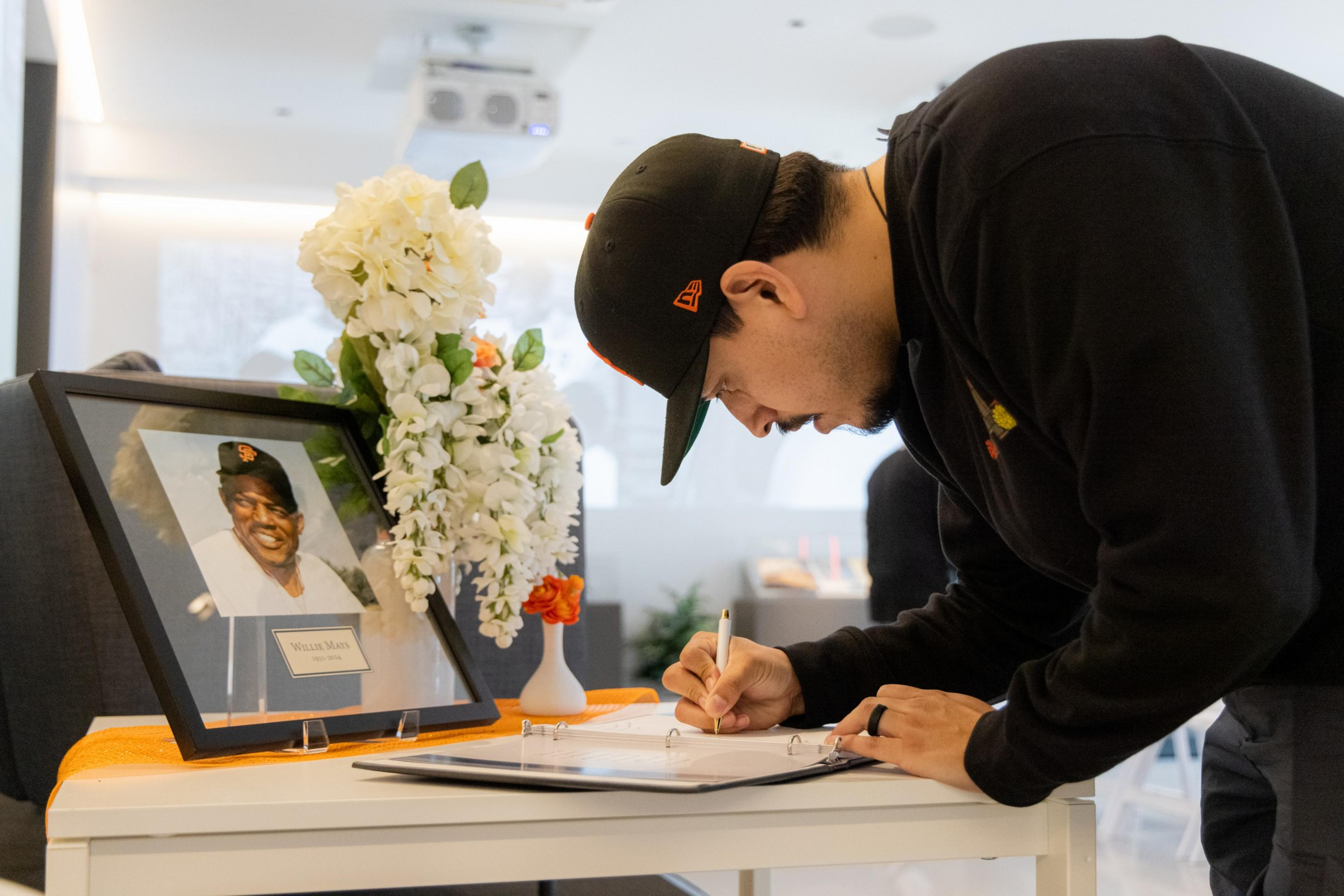A man wearing a black cap and sweater signs a guestbook on a small table. The table holds a framed photo of a smiling older man, alongside a bouquet of white flowers.