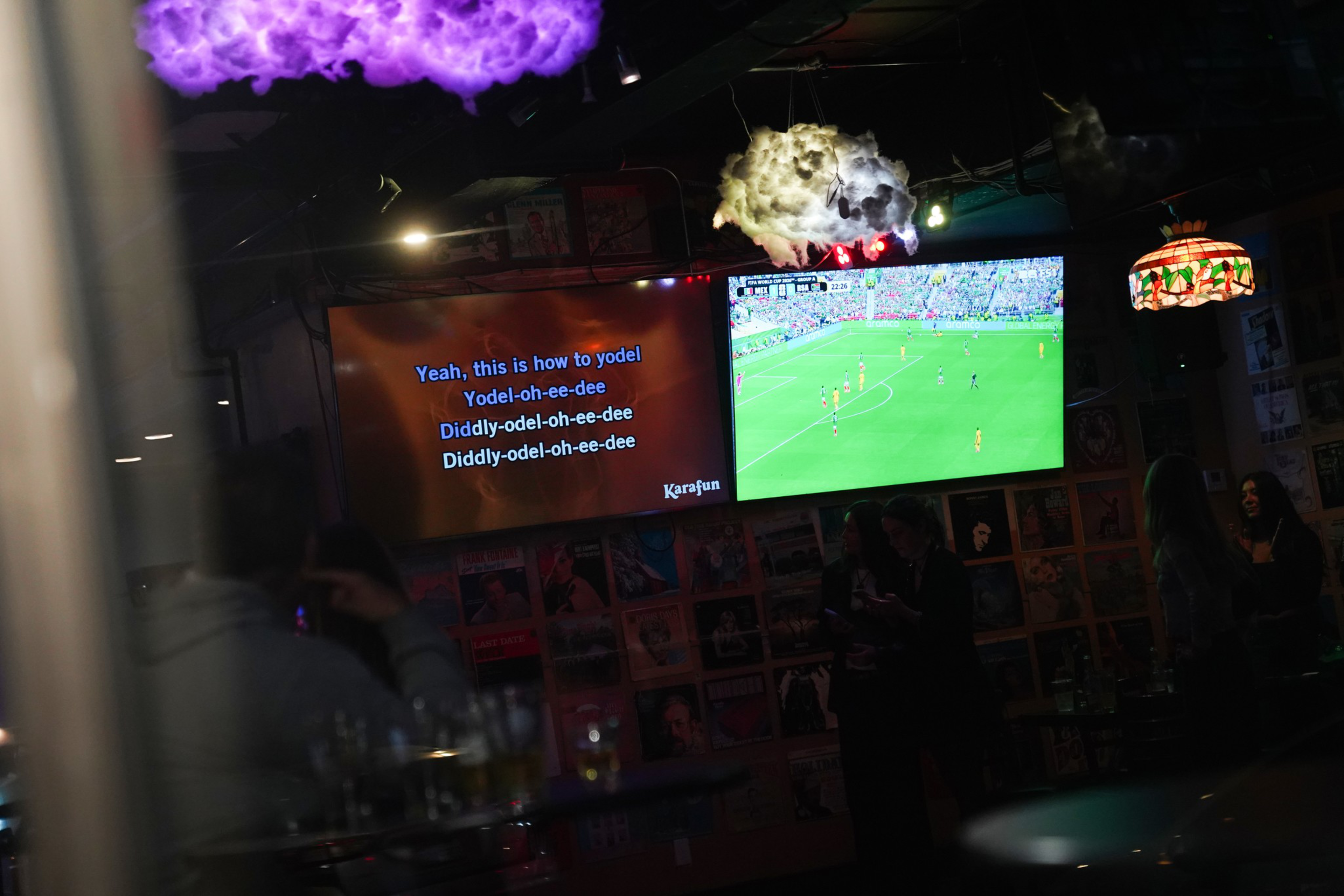 A karaoke screen shows “Yeah, this is how to yodel” lyrics beside a TV displaying a soccer match, with people standing and colorful cloud-shaped lights above.