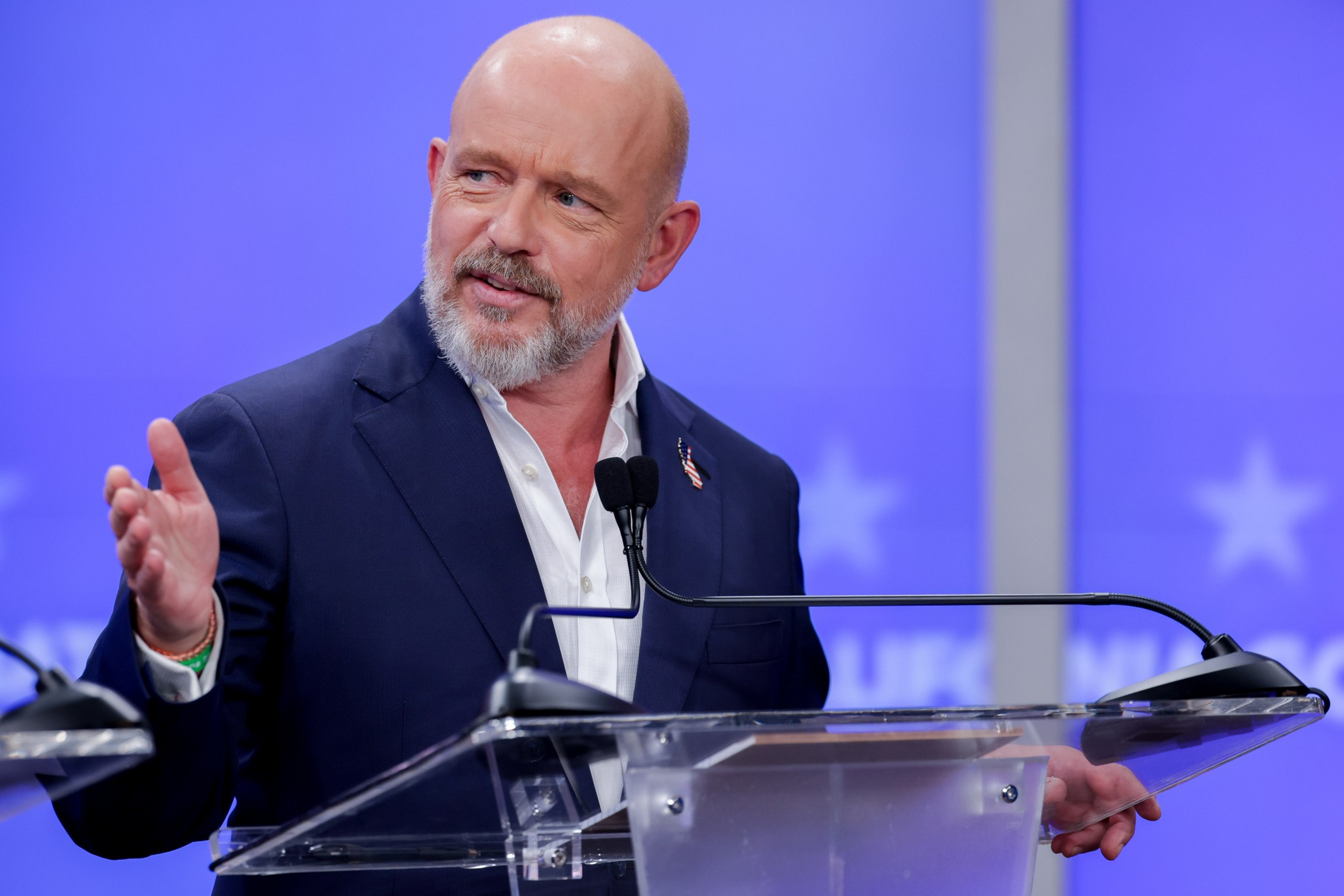 A bald man with a gray beard in a dark suit speaks at a clear podium with microphones, gesturing with his left hand against a blue background.