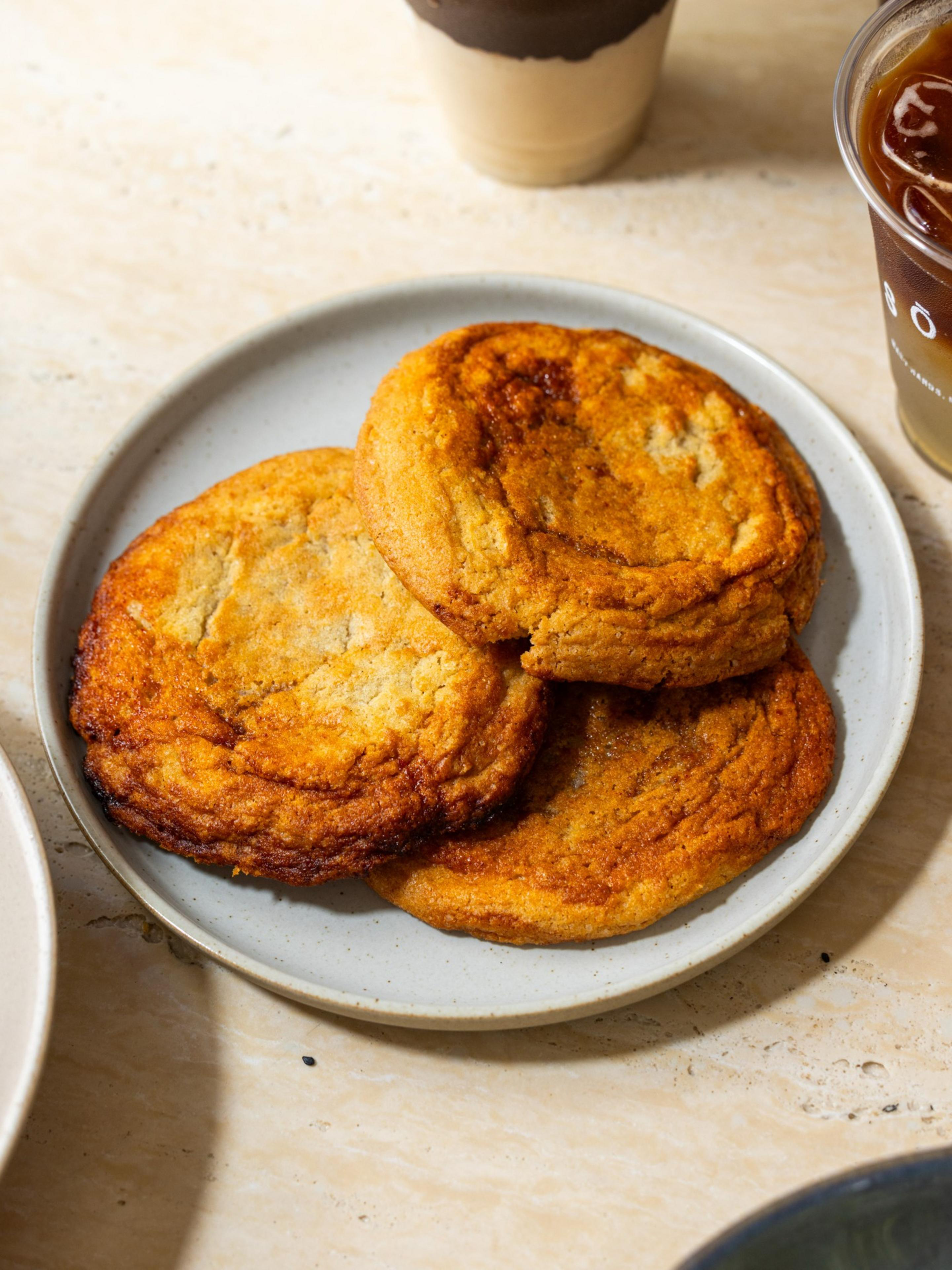 Three golden-brown, slightly crispy cookies rest on a simple gray plate, with a blurred iced drink and cup in the background on a light-colored surface.