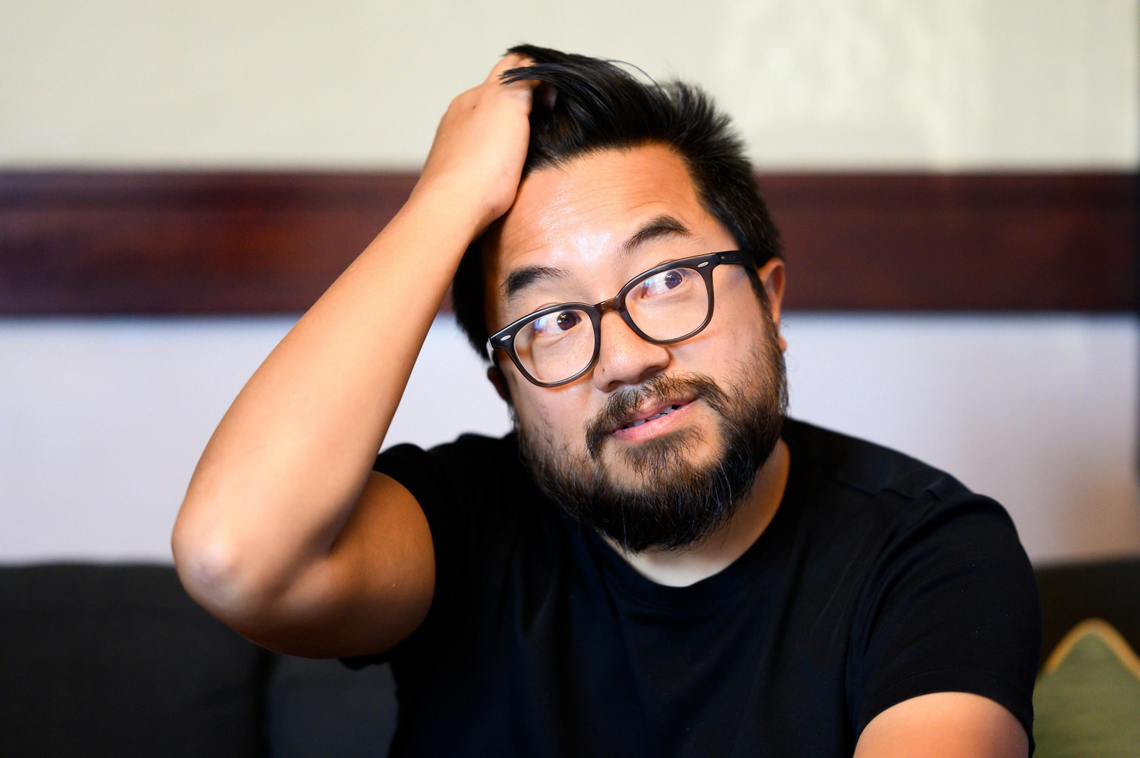 A bespectacled man in a black shirt, hand in hair, looks at the camera with a slight smile.