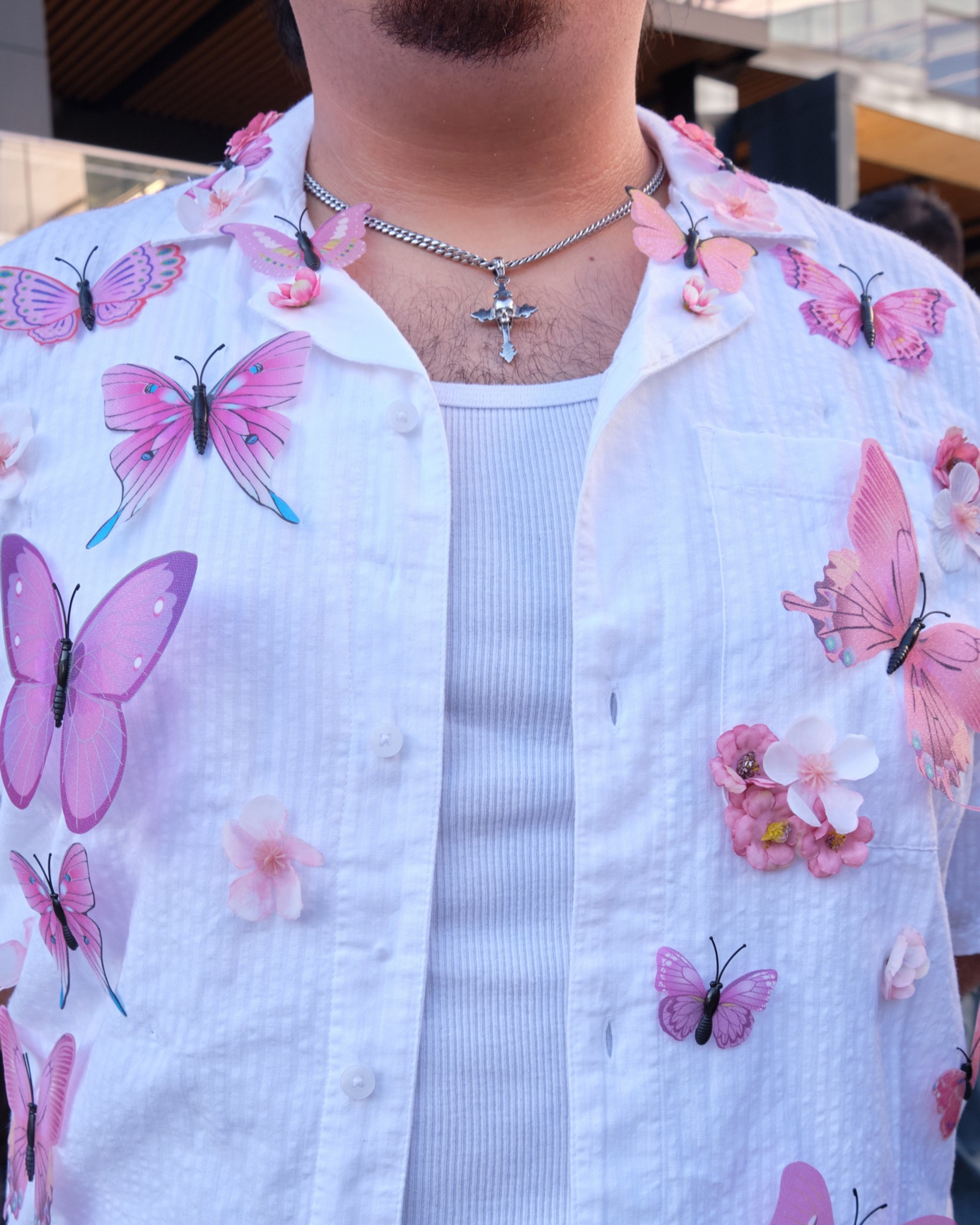 A person wears a white shirt decorated with pink butterflies and flowers over a white ribbed tank top, with a silver chain necklace featuring a cross pendant.