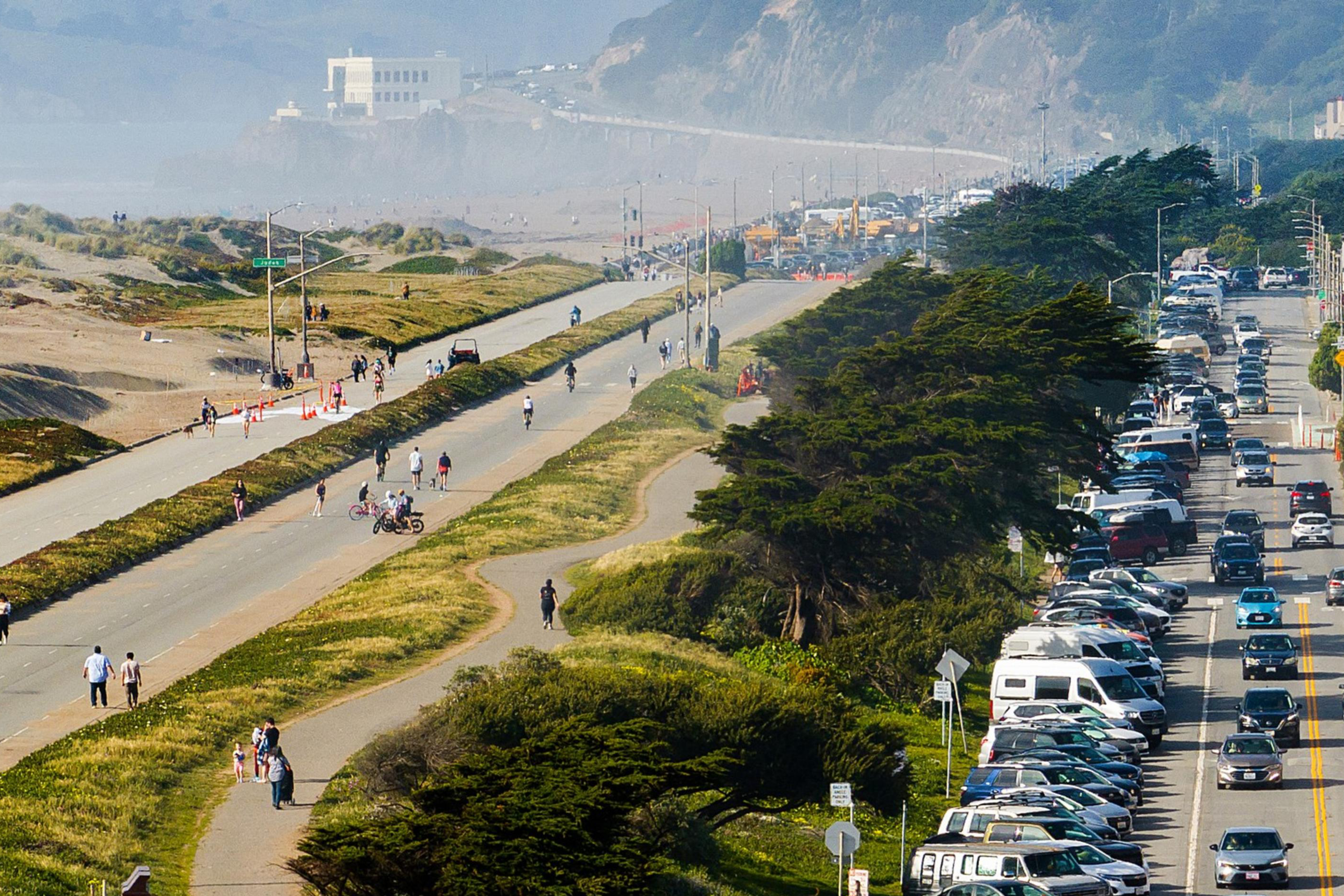 A busy road with heavy traffic and parked cars is adjacent to a separate pedestrian path and bike lane. People are walking and cycling under clear skies.