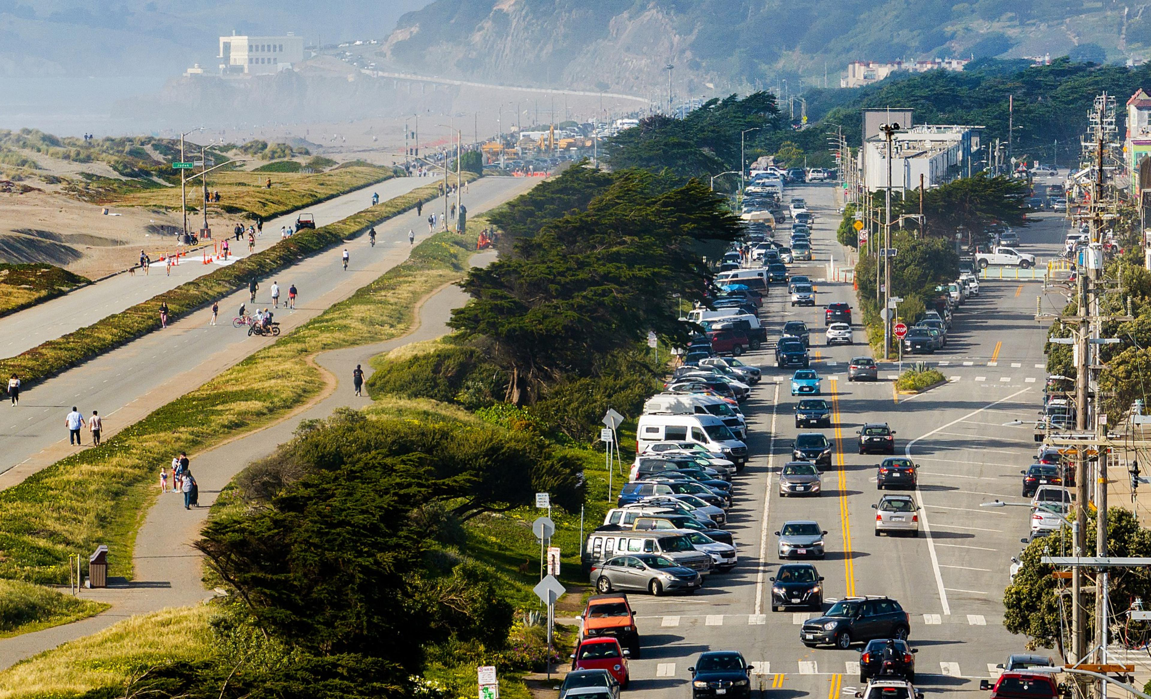 A busy road with heavy traffic and parked cars is adjacent to a separate pedestrian path and bike lane. People are walking and cycling under clear skies.