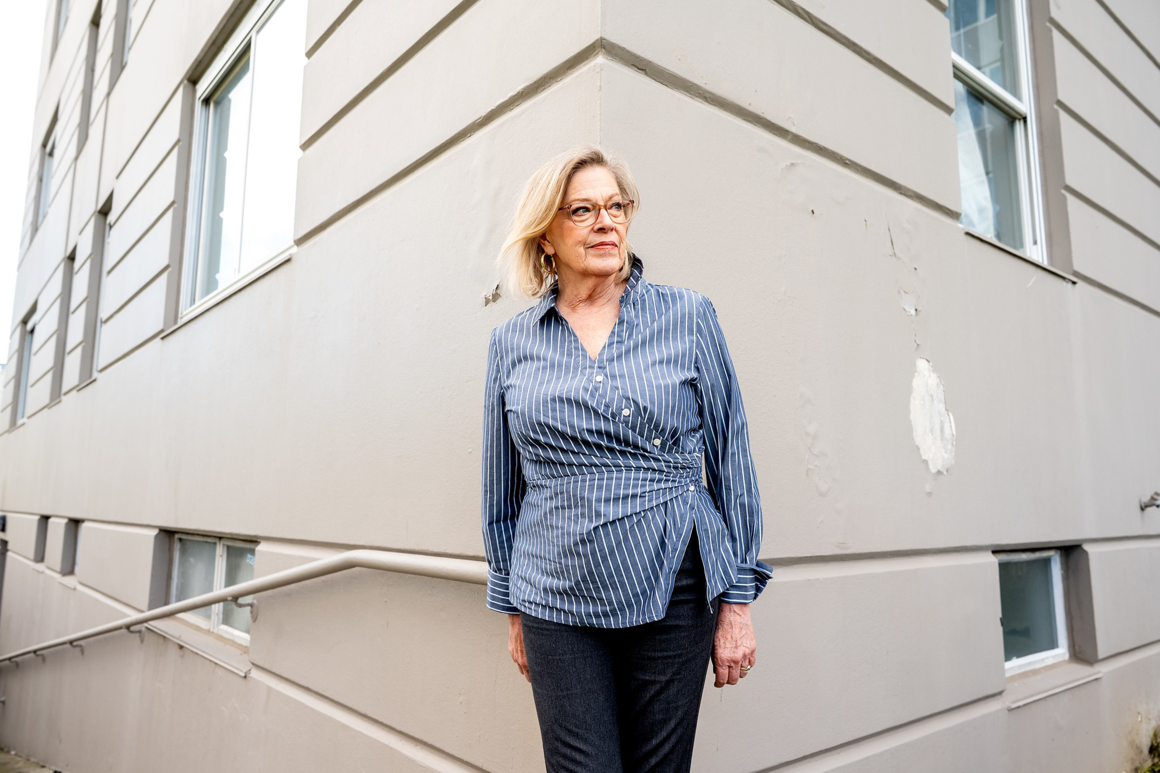A woman with blonde hair and glasses stands near a corner of a beige building, wearing a blue striped blouse and dark pants, looking to the side.