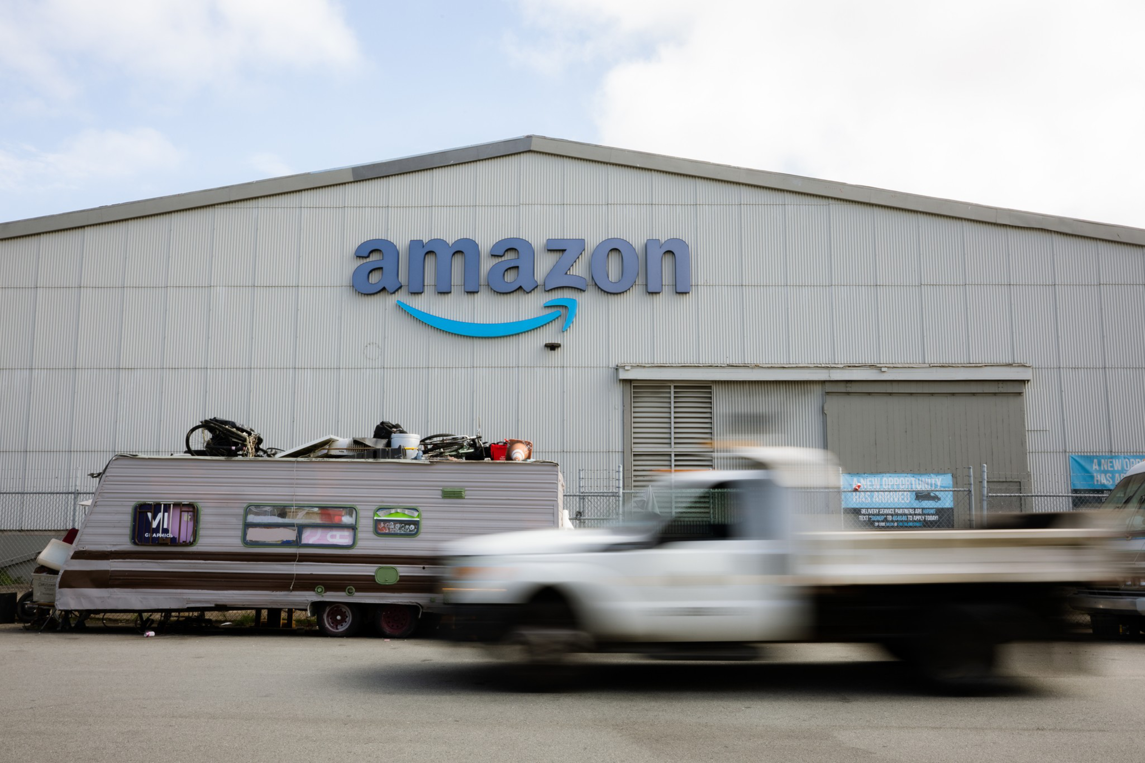 A white Amazon warehouse building with a large logo has a worn trailer parked in front, while a white pickup truck drives past blurred in motion.