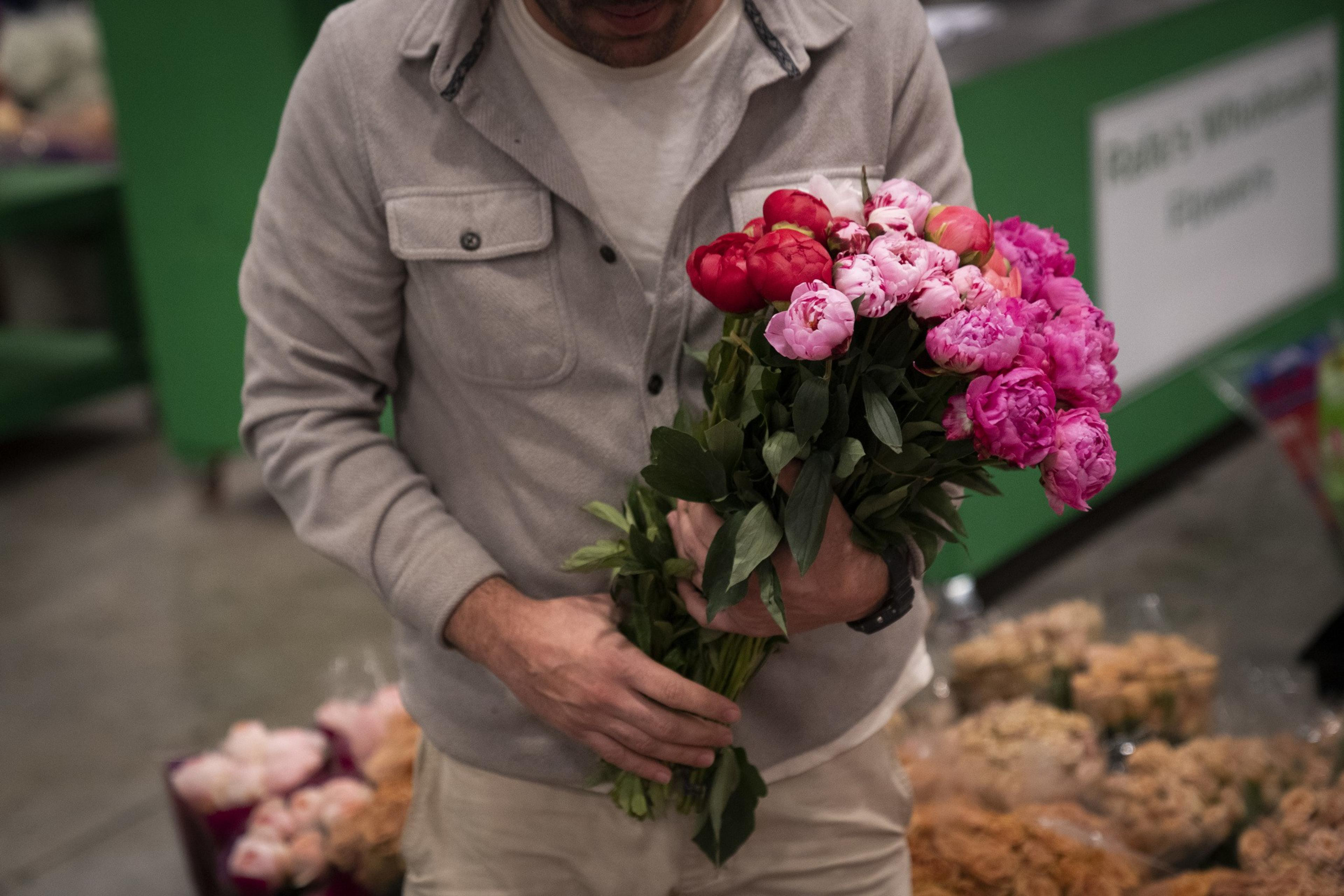 A person in a gray jacket holds a bouquet of pink and red peonies, standing in a market setting with colorful flower bouquets in the background.