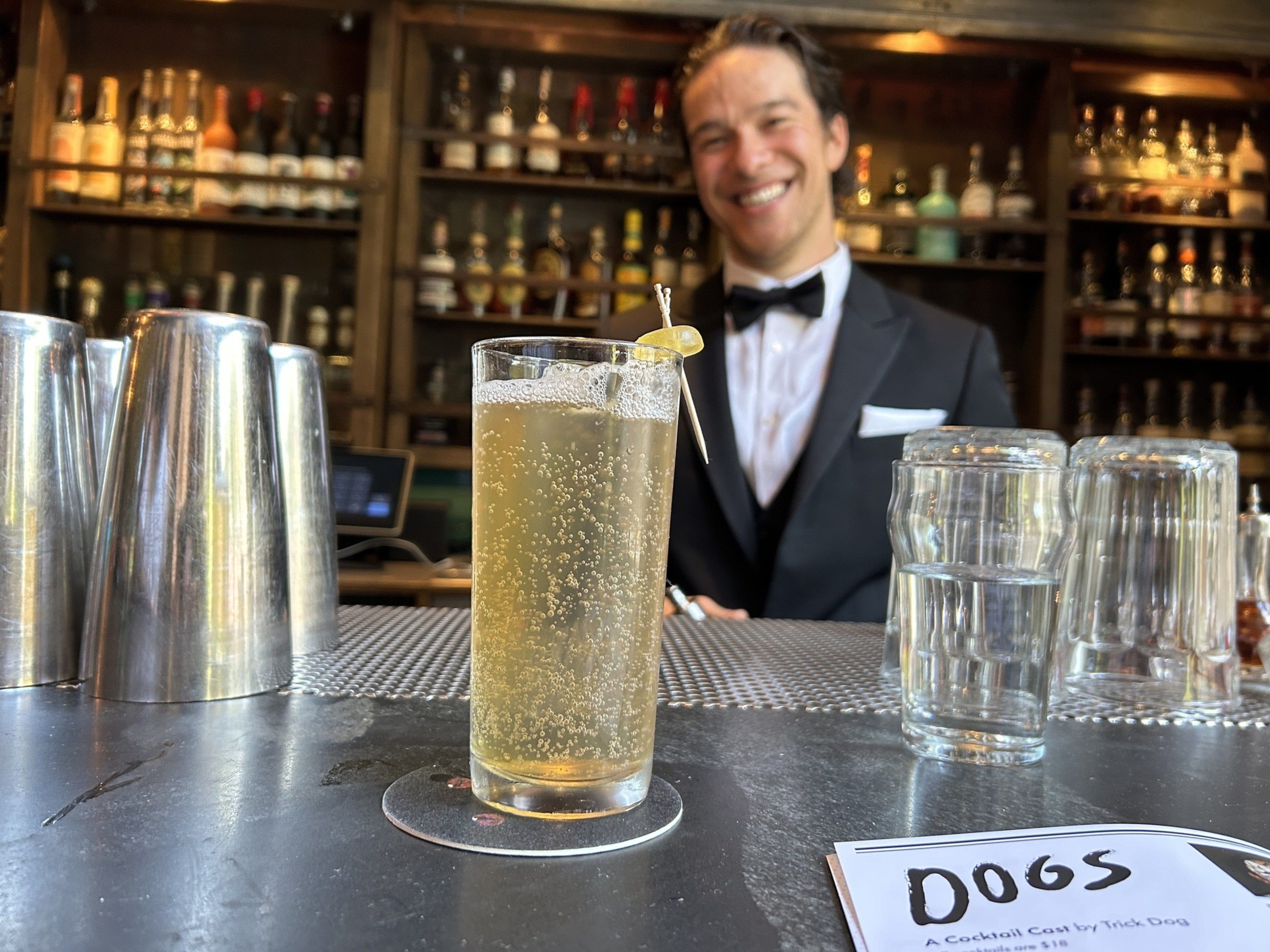 A smiling bartender in a tuxedo is behind the bar. A bubbly cocktail garnished with a lemon twist is on the counter next to stacked glasses.