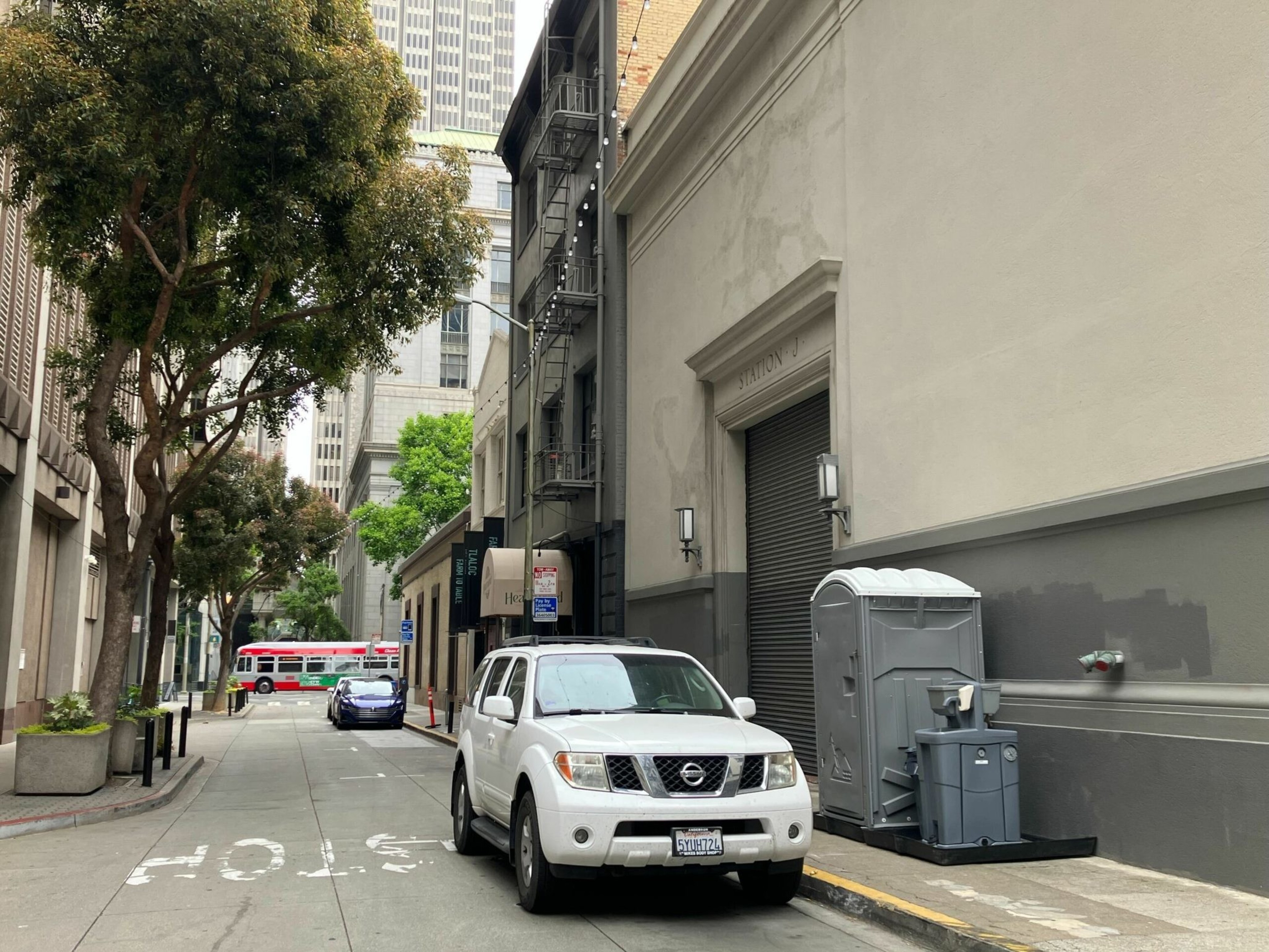 A white Nissan SUV is parked on a narrow city street next to a gray portable toilet and a trash bin, with trees and a red bus visible in the background.