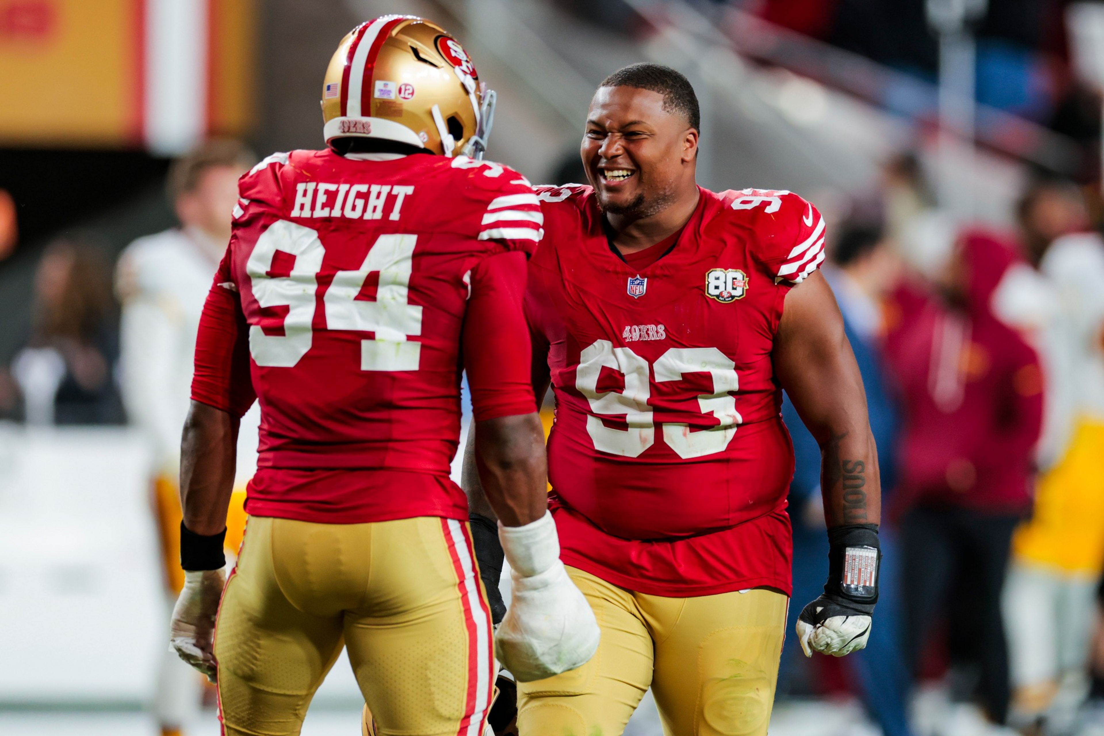Two San Francisco 49ers football players, wearing red jerseys numbered 94 and 93, share a joyful moment on the field.