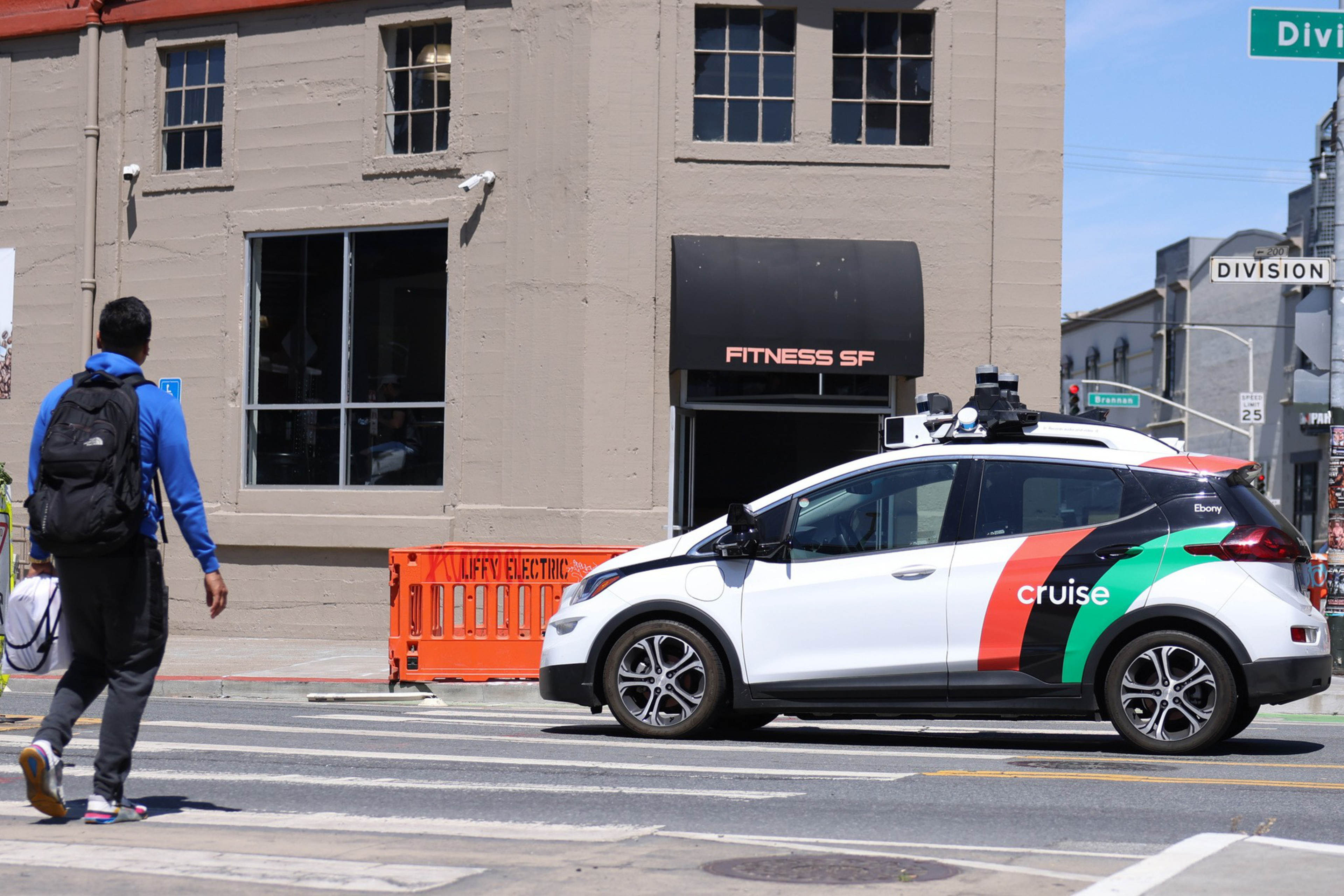 A white Cruise autonomous car with red and green stripes waits at a crosswalk while a pedestrian wearing a blue hoodie and black backpack crosses the street.