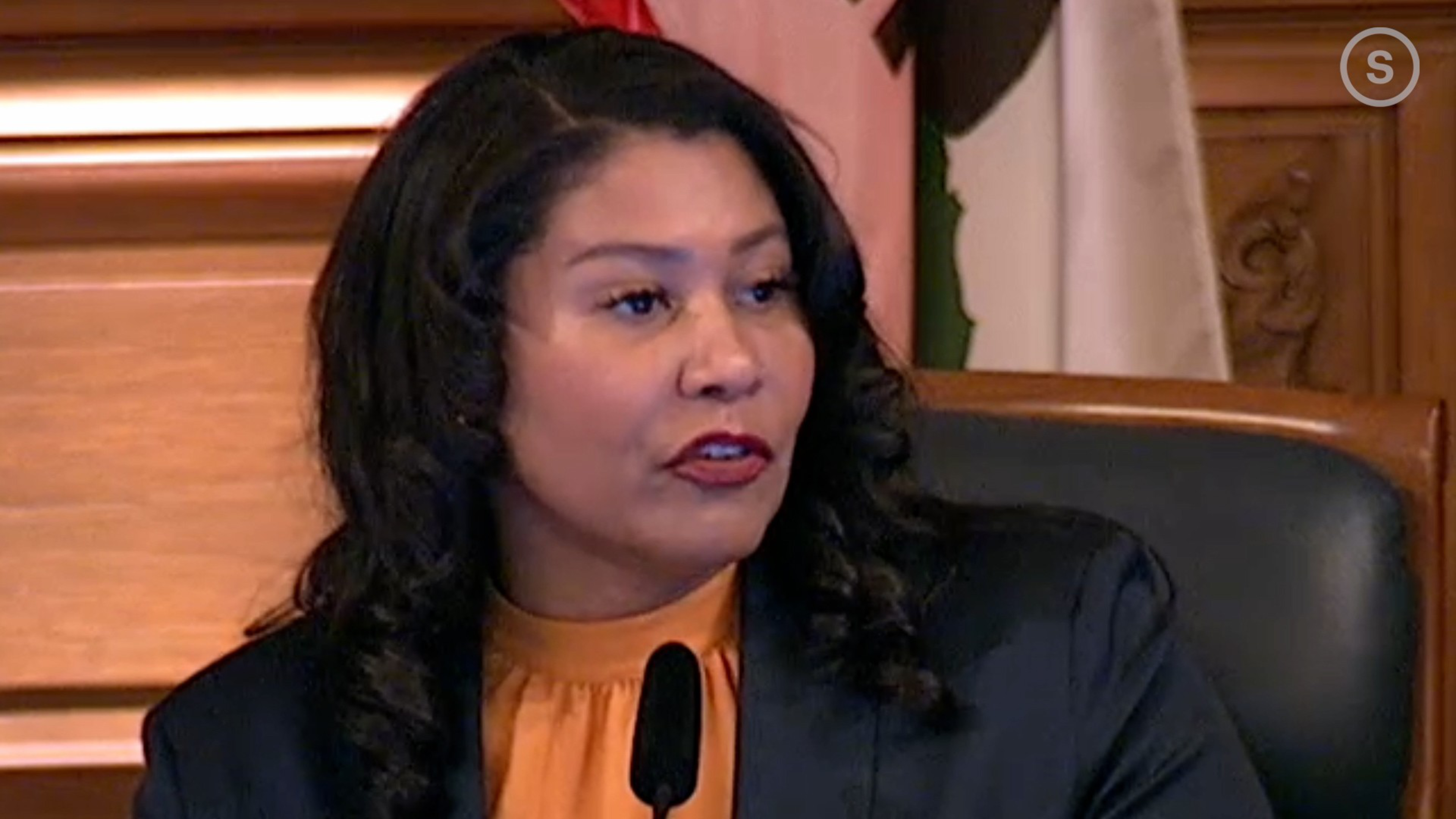 A woman with long curly hair wears a black blazer and orange blouse, speaking in front of a microphone with wooden paneling behind her.