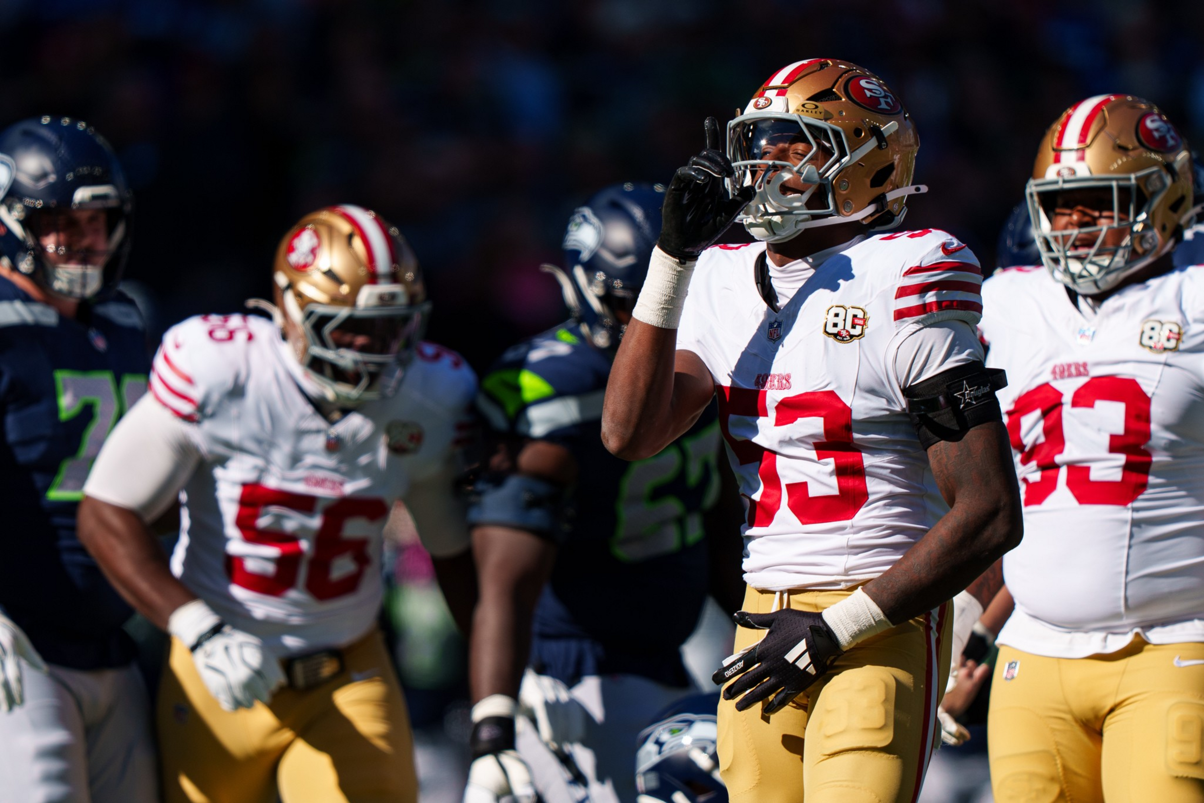 A San Francisco 49ers player in a white and red jersey raises his hand near his helmet during a game against Seattle Seahawks players in dark blue and green jerseys.