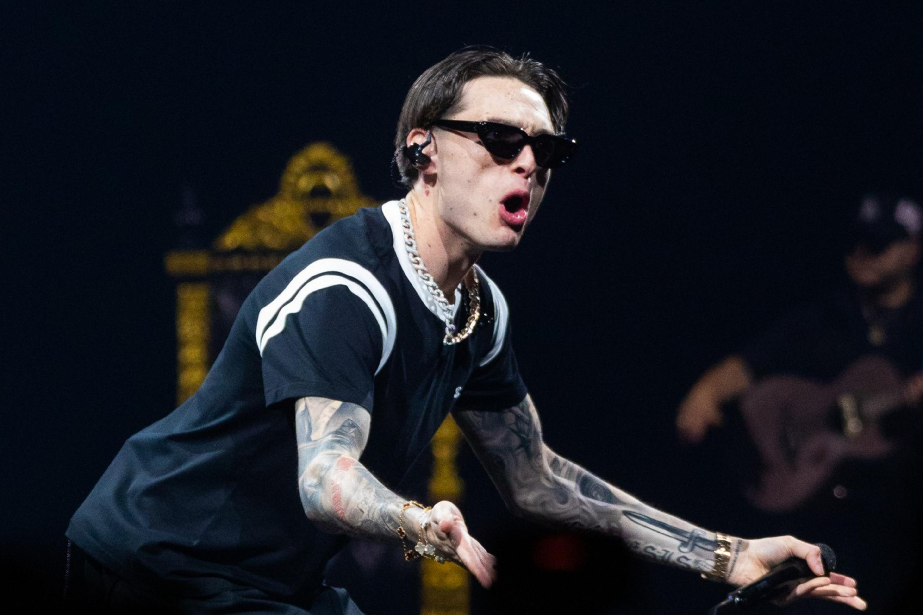 A tattooed man, wearing sunglasses, a black shirt, and a gold chain, performs energetically on stage, singing into a mic with an intense expression. A band member is in the background.