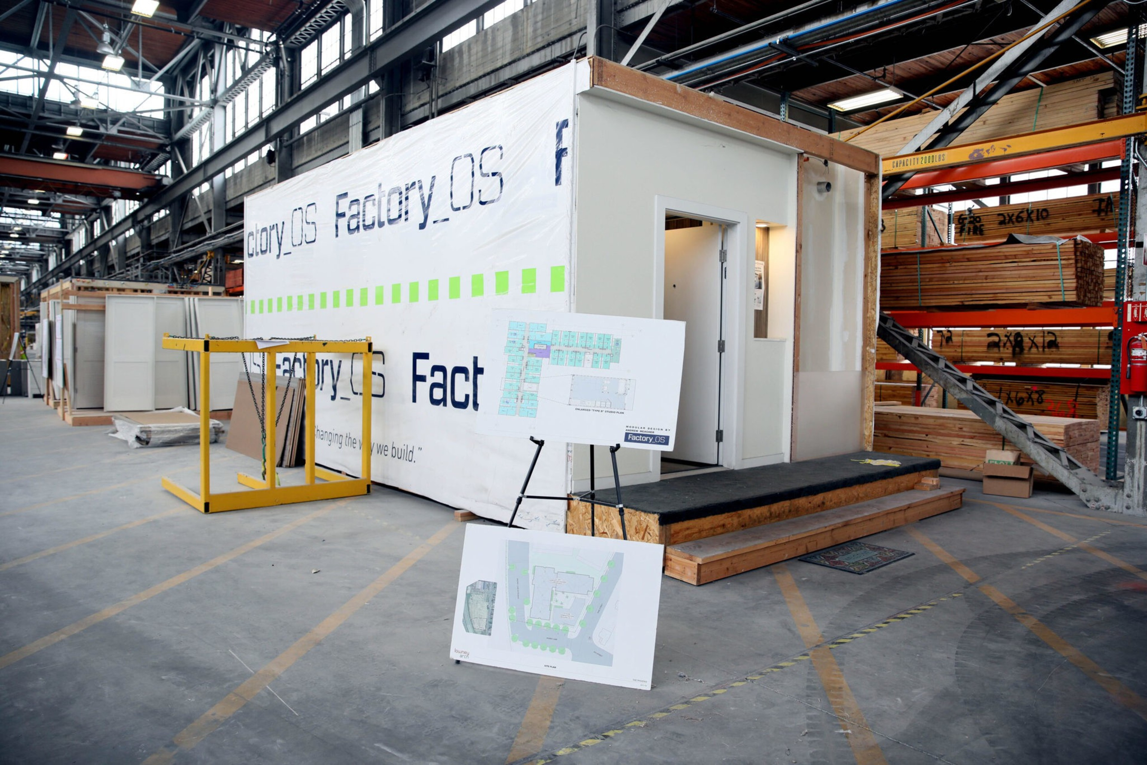 A prefab modular building unit sits inside a warehouse with construction plans displayed on boards nearby and lumber stacked on shelves.