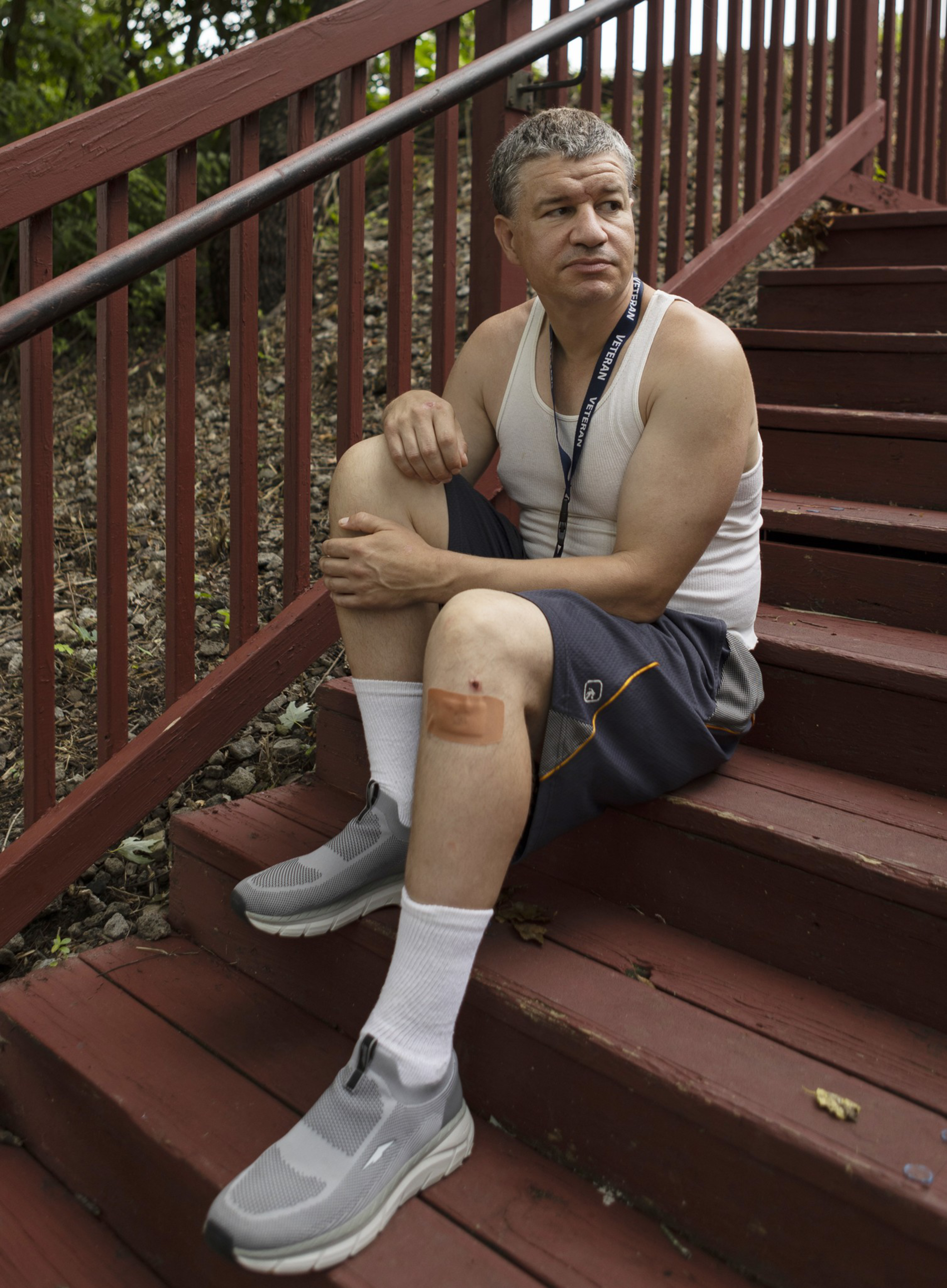 A man in a white tank top and gray shorts sits on red wooden stairs outdoors, resting his arm on his knee, with a visible bandage on his shin.
