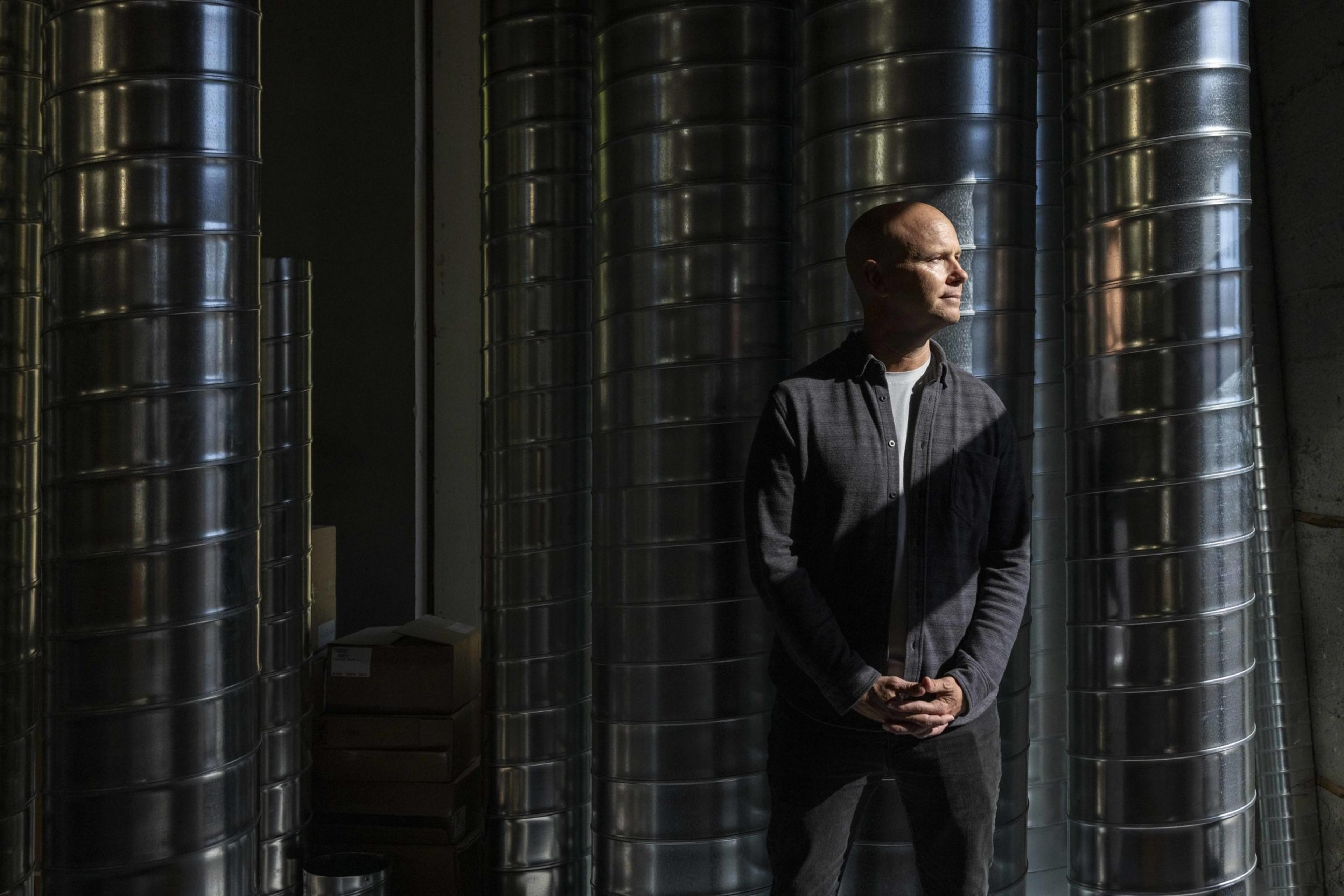 A person stands in a dimly lit room surrounded by large metal ducts. They are dressed in a dark shirt and pants, with light casting shadows across their face.