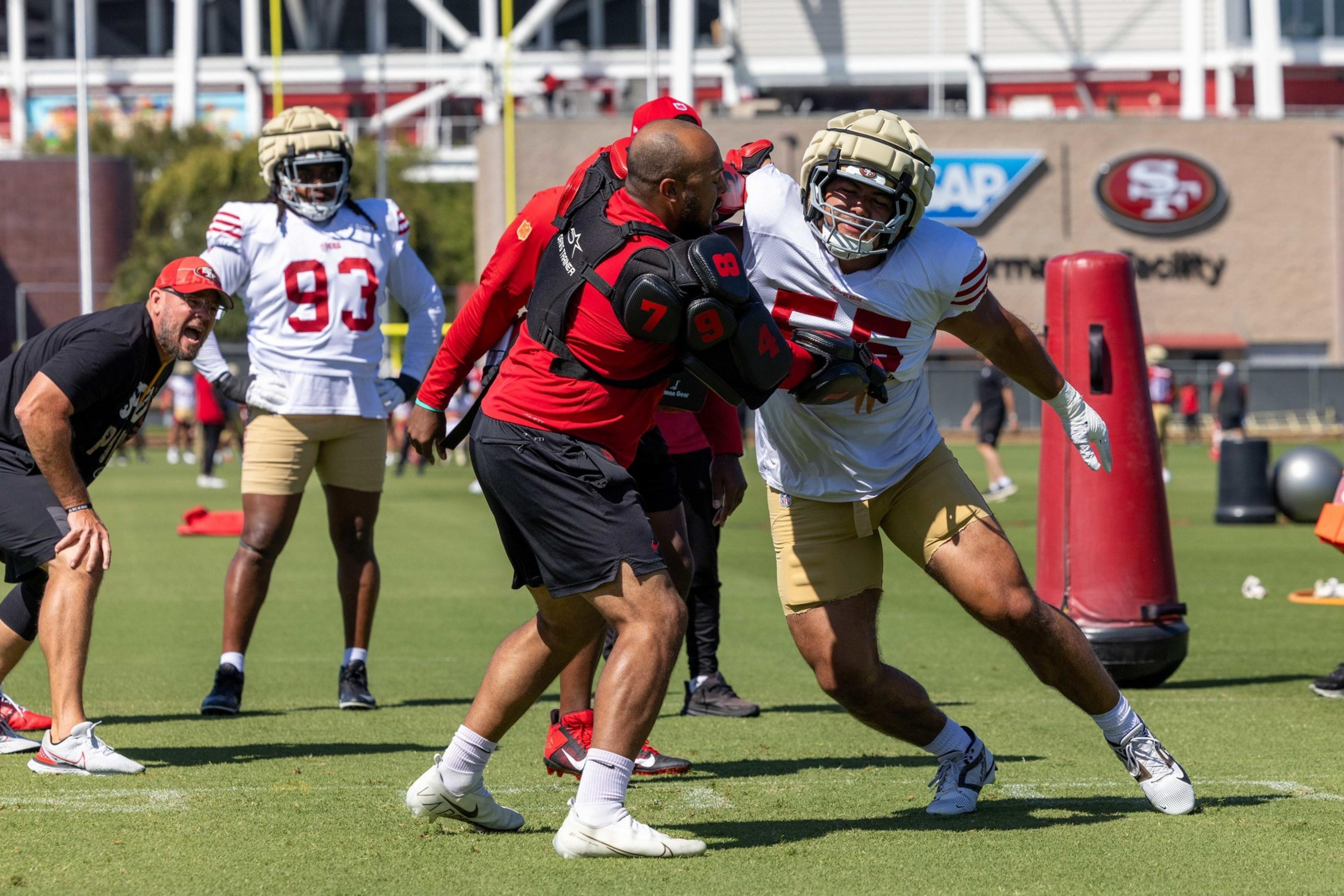 A football player wearing a white jersey and gold shorts pushes against a coach in a red padded vest during a practice drill.