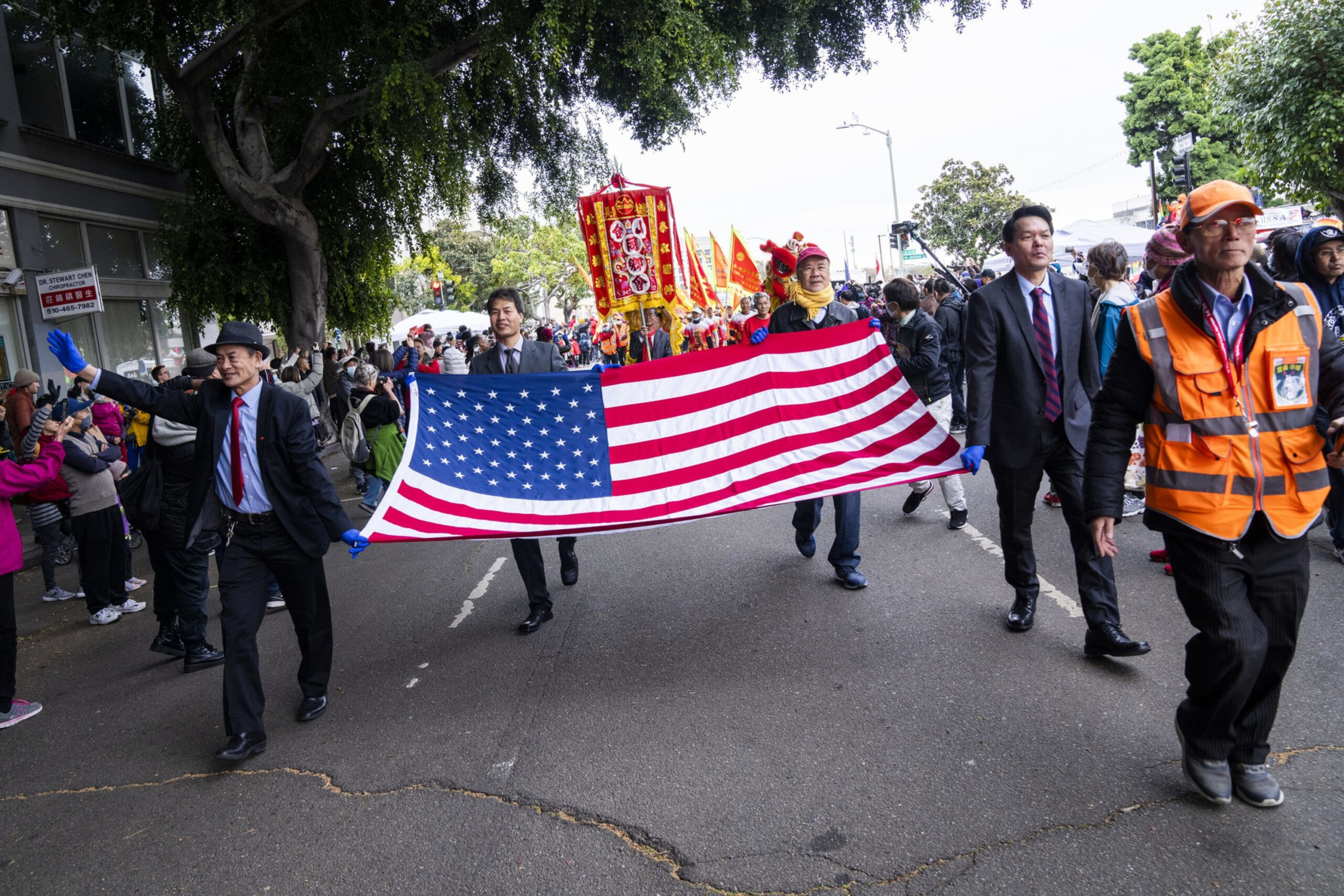 Four men in suits and gloves hold a large American flag horizontally during a street parade with colorful banners and a crowd watching.