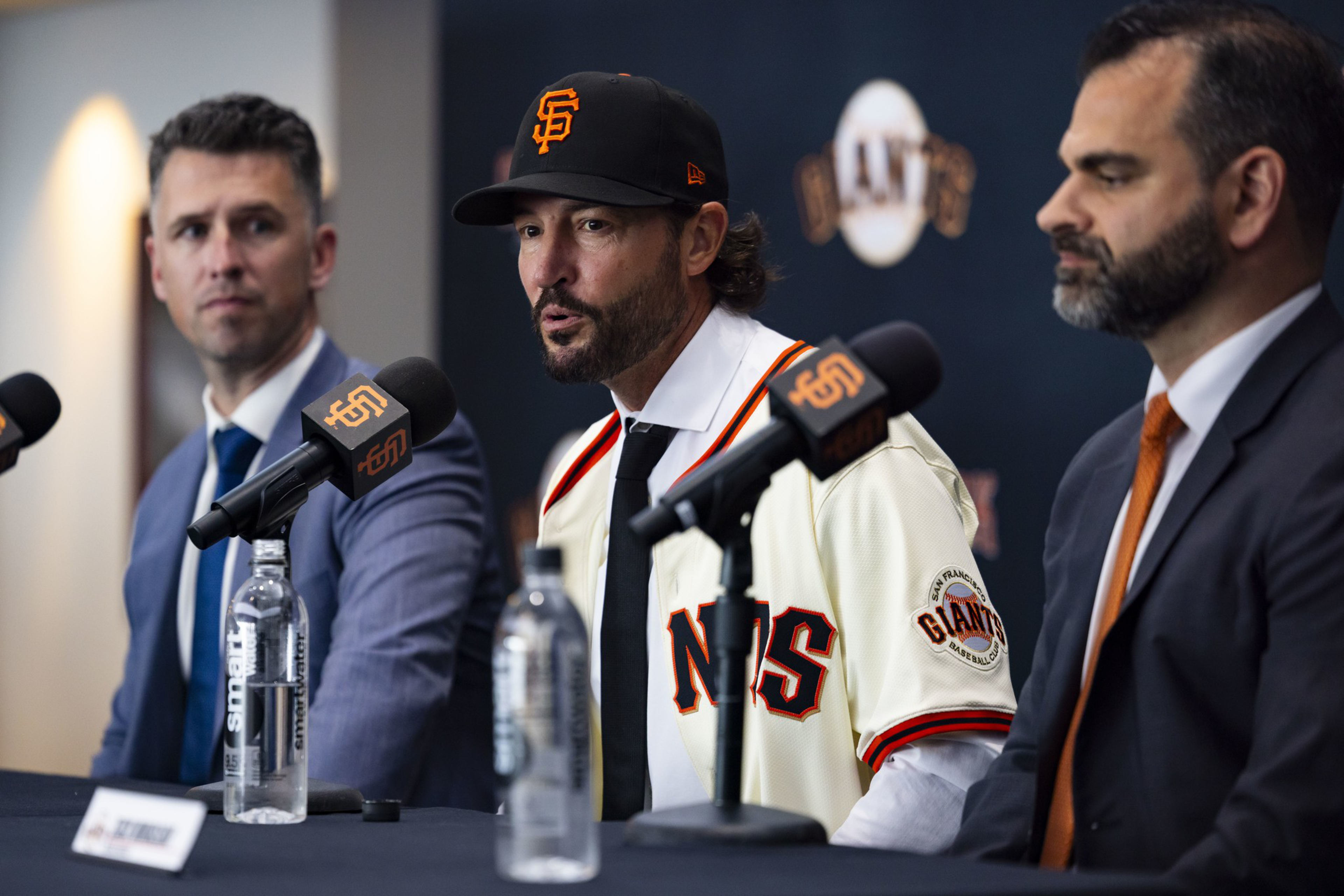 A man in a San Francisco Giants jersey and cap speaks into a microphone, flanked by two suited men at a press conference table.