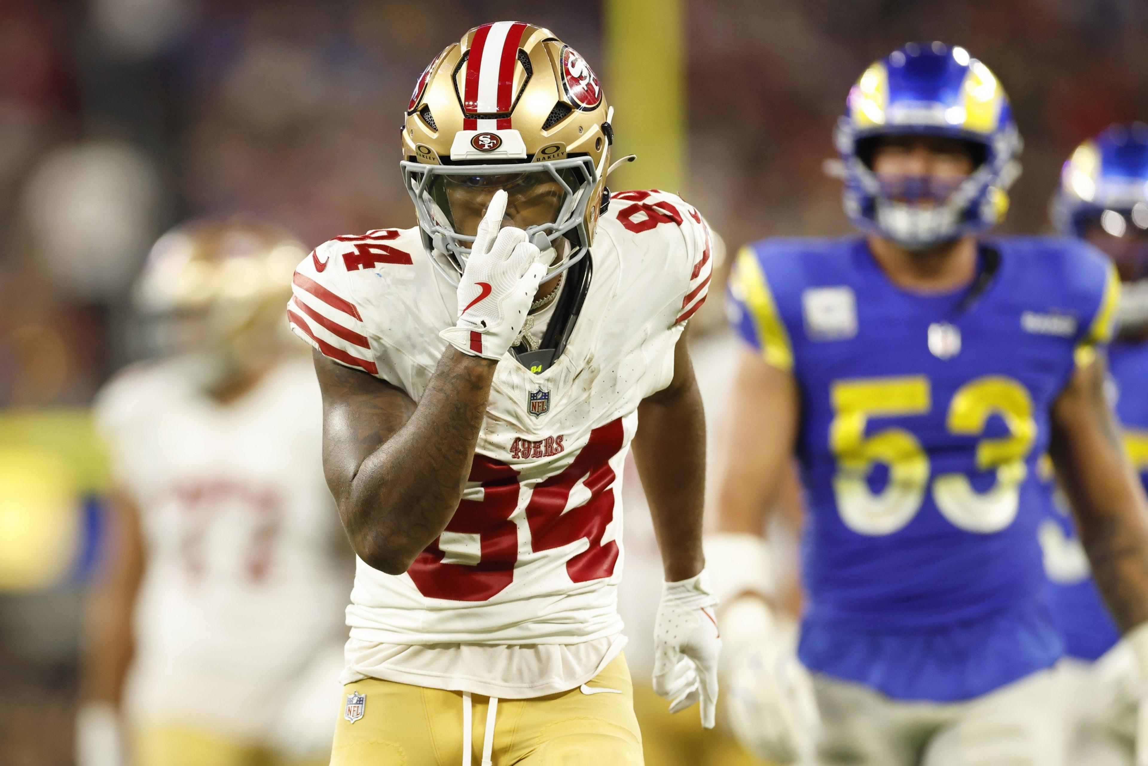 A 49ers player, number 84, points to his helmet while running, with two Rams players, including number 53, in the blurred background.