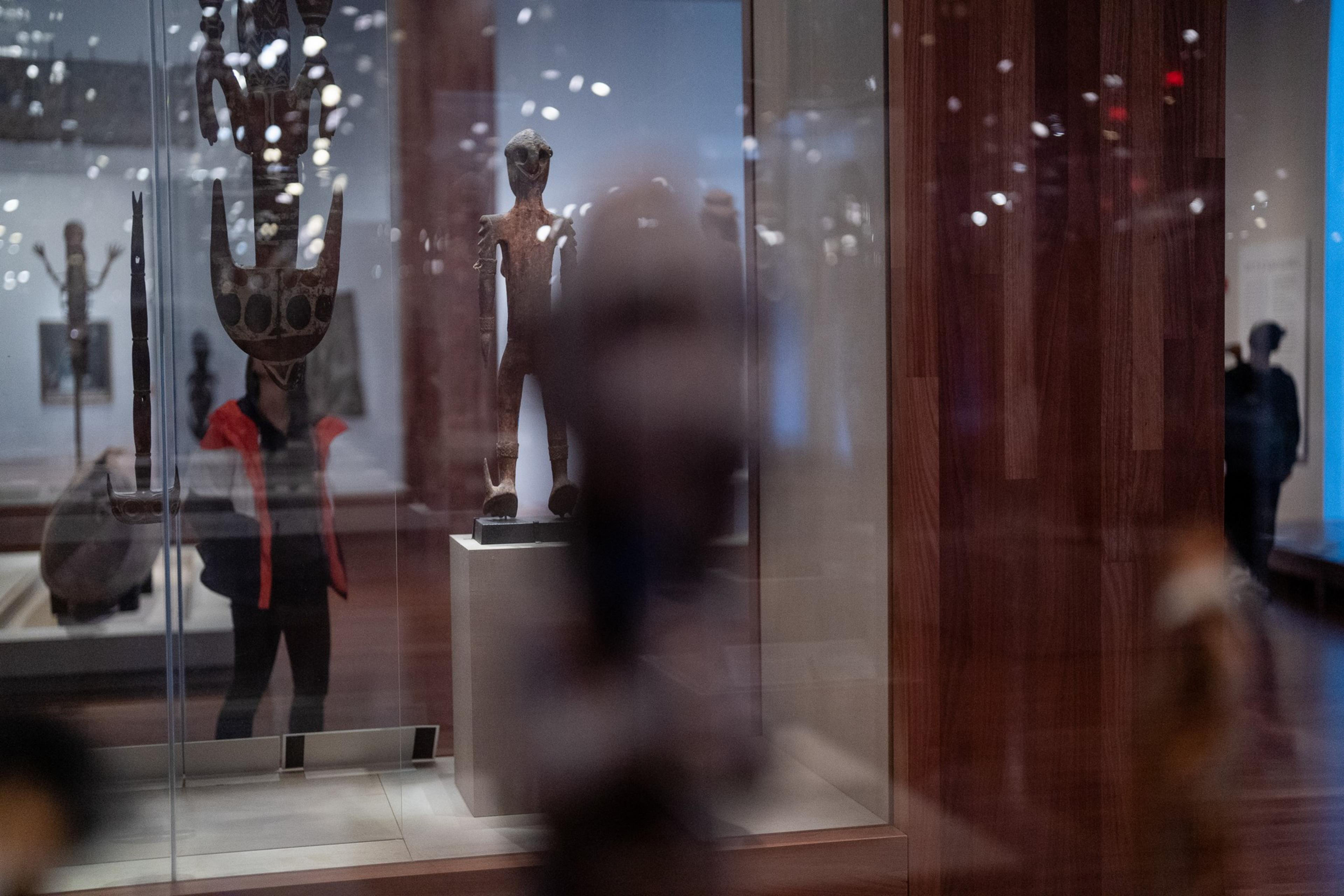 The image shows a museum exhibit with wooden sculptures displayed in glass cases. Reflections of visitors are visible in the glass, creating a layered, abstract effect.