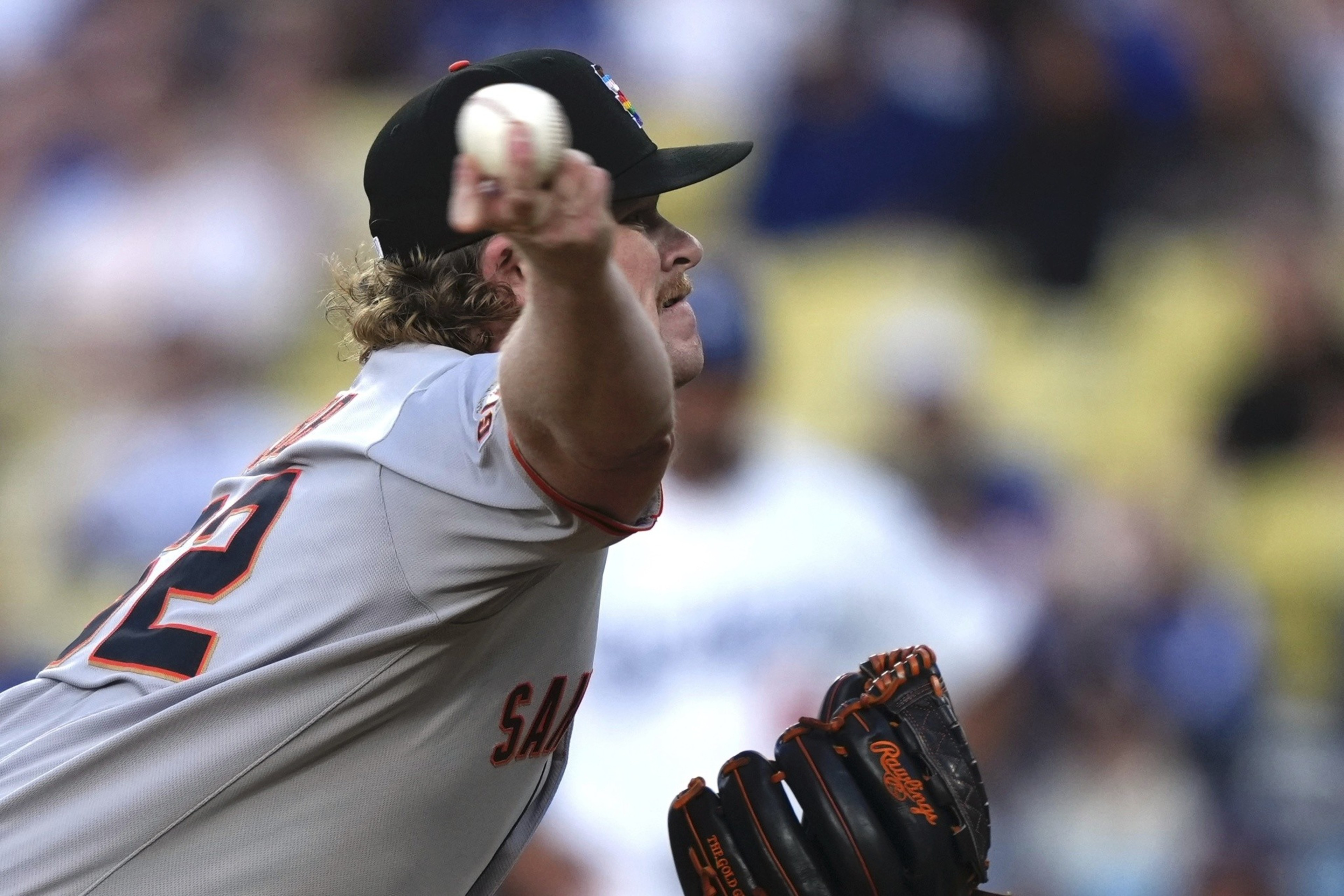 A baseball pitcher in a grey jersey throws a ball, his glove labeled &quot;Rawlings.&quot; The focus is on his arm and the ball, with the crowd blurred in the background.