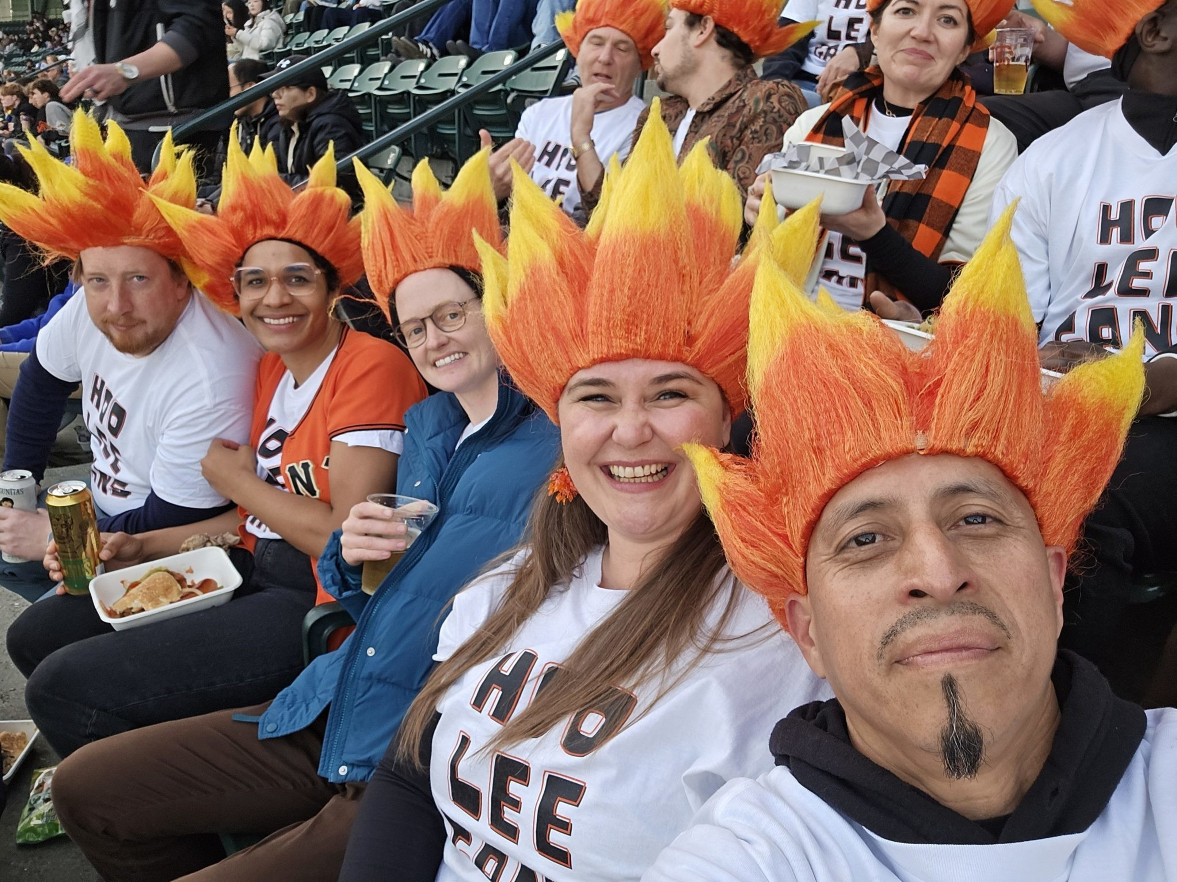A group of smiling fans wearing bright orange and yellow flame wigs and matching &quot;Hoo Lee&quot; shirts, sitting in stadium seats with food and drinks.