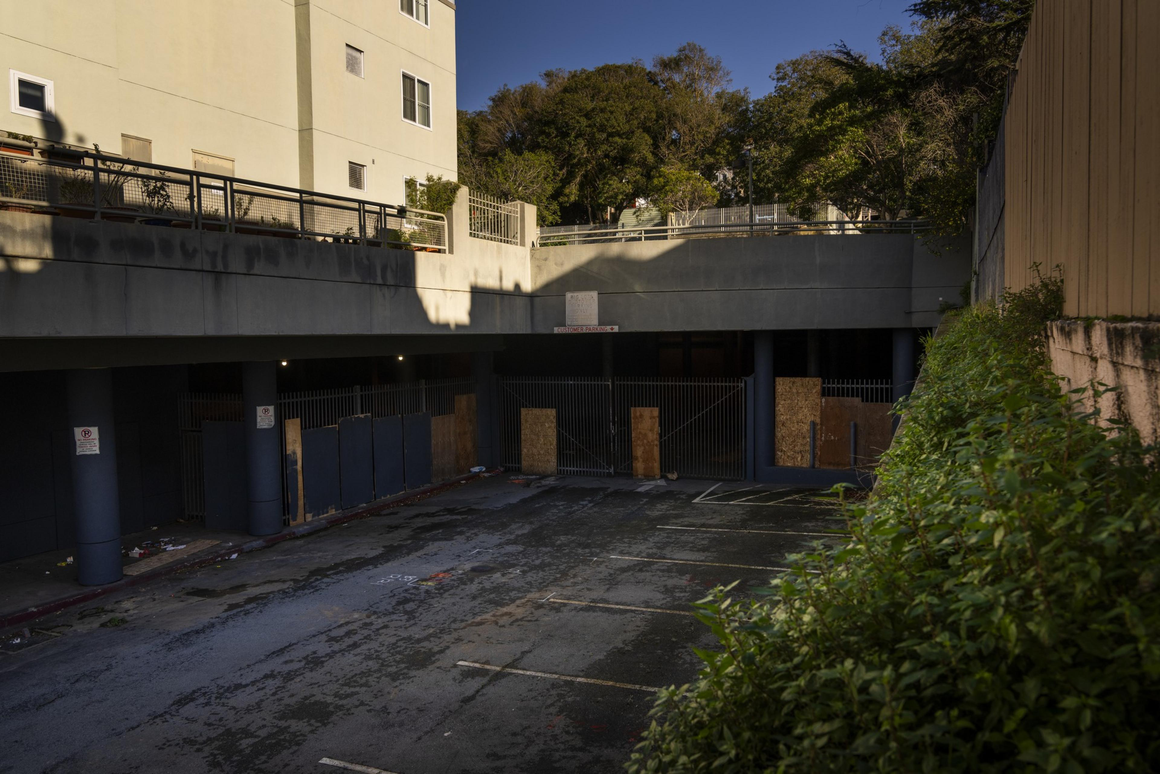 An empty parking lot with boarded-up entrances, adjacent buildings, and overgrown foliage under a clear sky.
