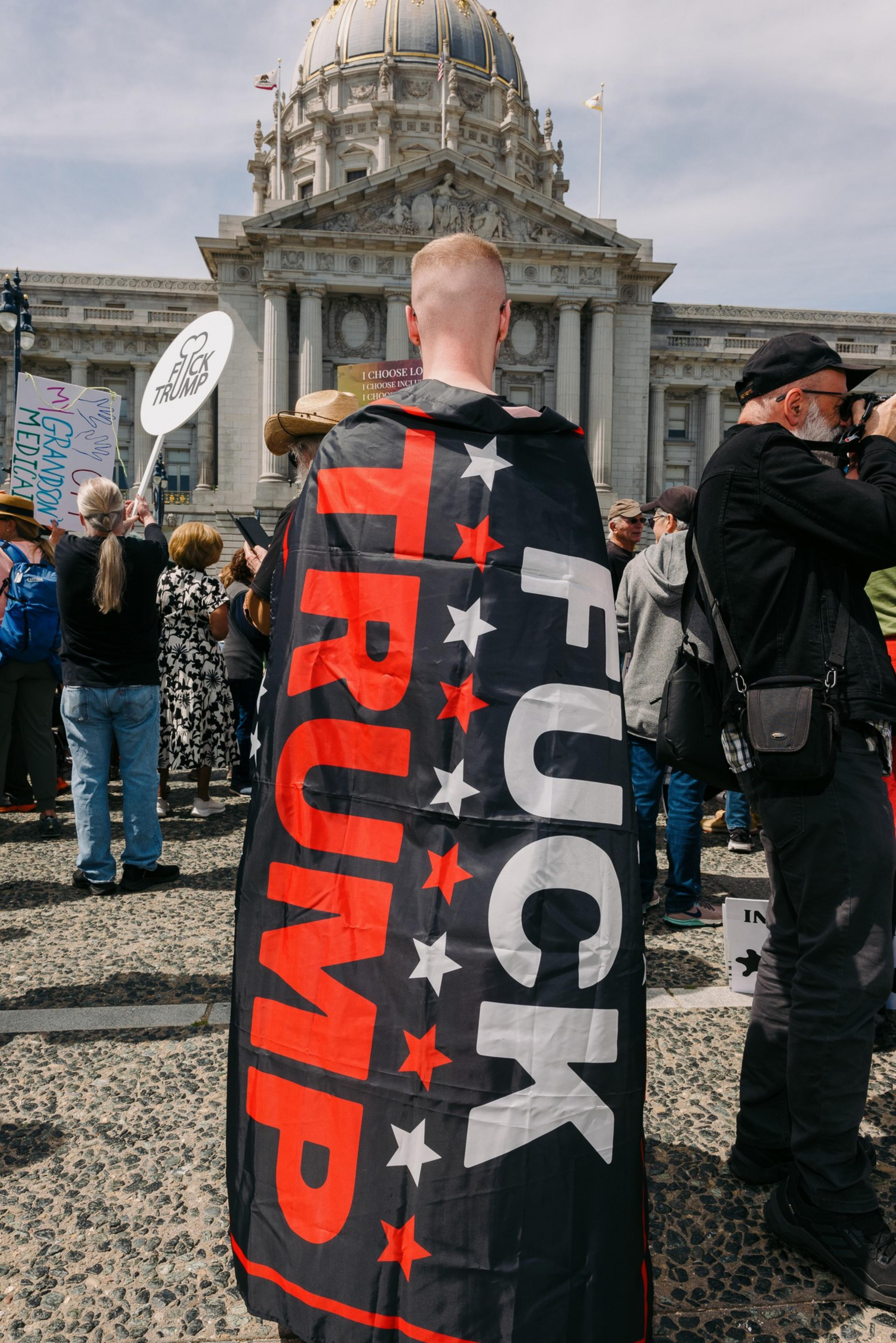 A person wrapped in a &quot;F*** Trump&quot; flag stands in a crowd at a protest outside a domed government building. Other signs and people are visible.