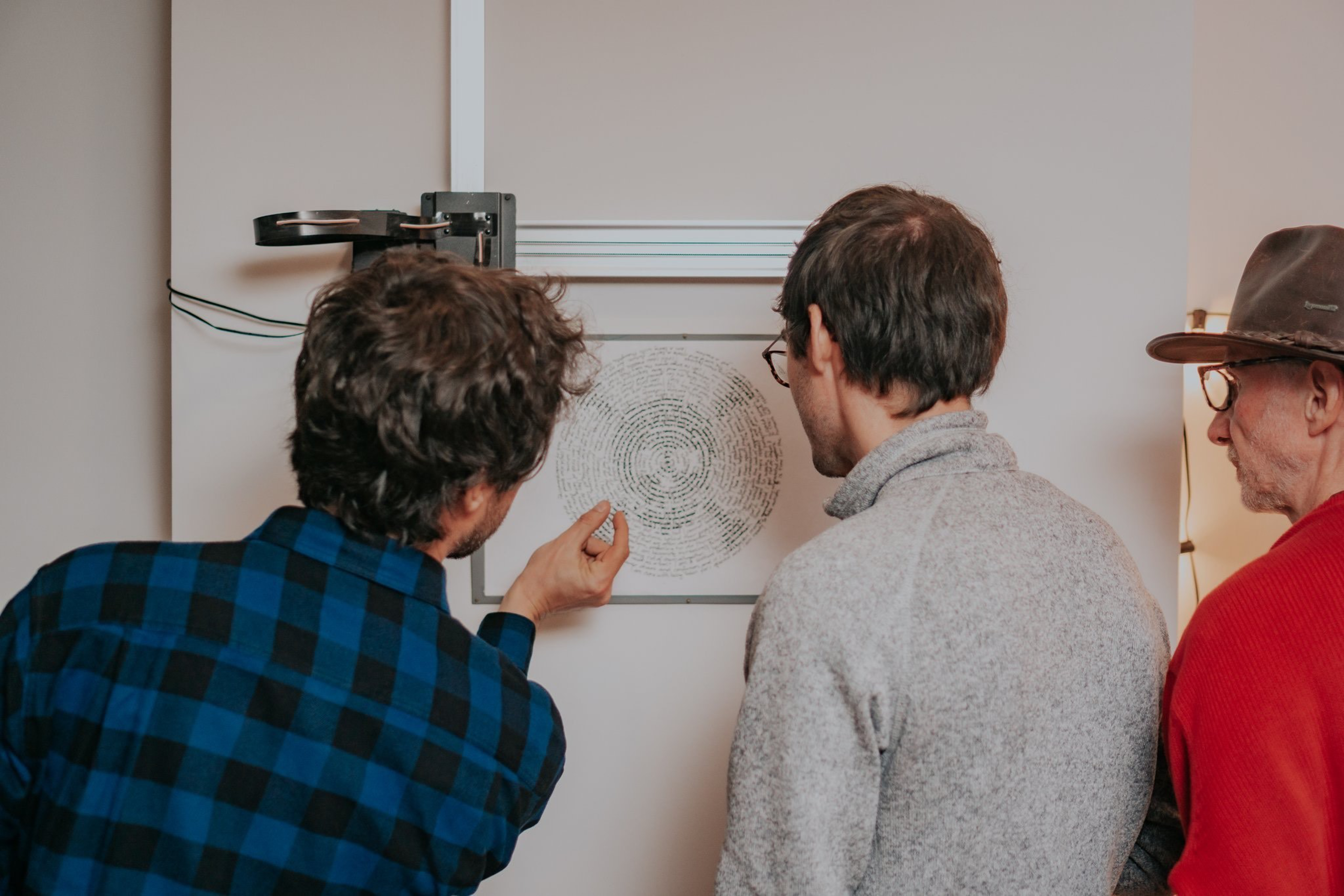 Three men closely examine a circular piece of paper with dense spiral writing mounted on a wall under a magnifying glass.