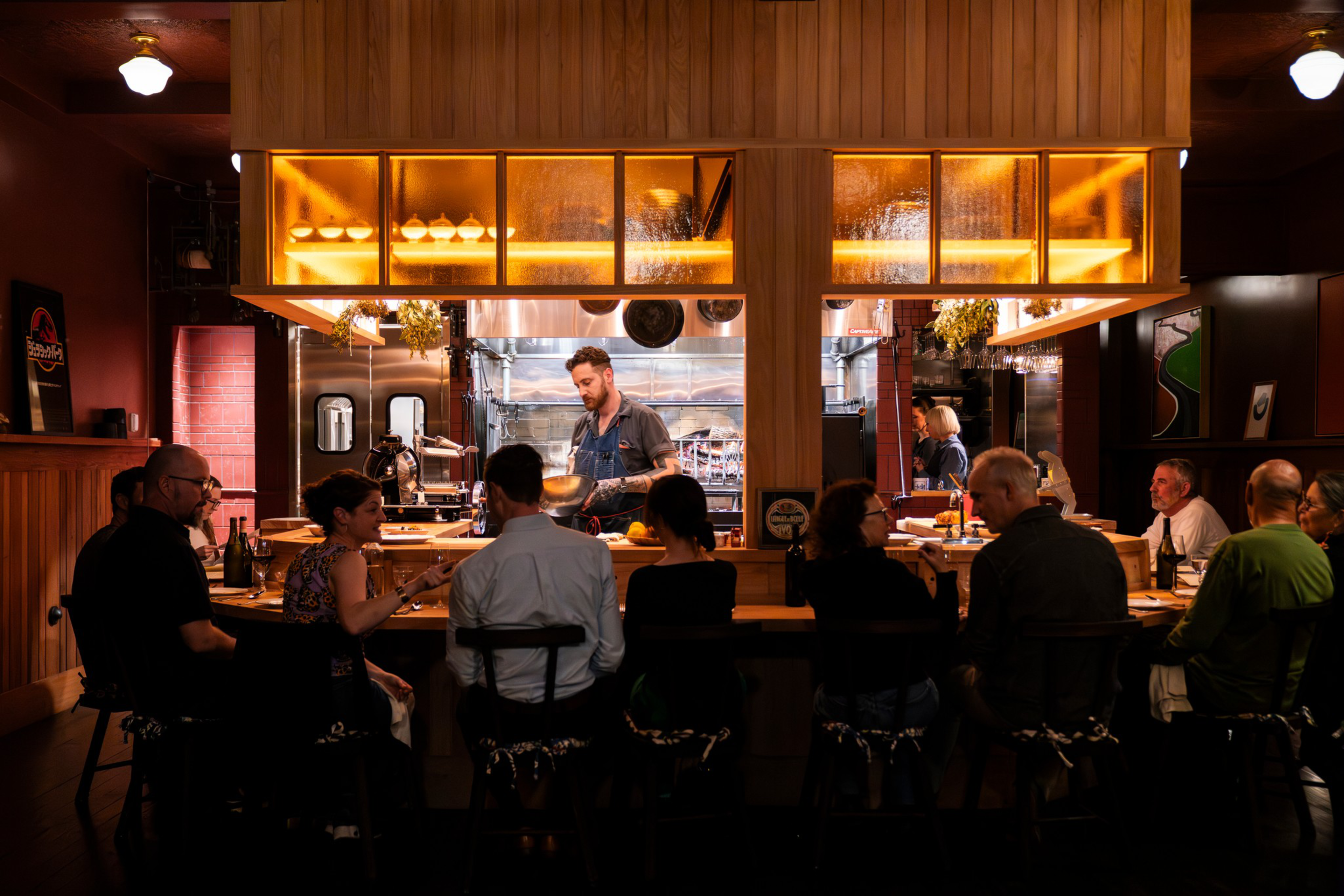 A chef prepares food behind a warm-lit counter while eight people sit on stools, facing the kitchen in a cozy restaurant setting.